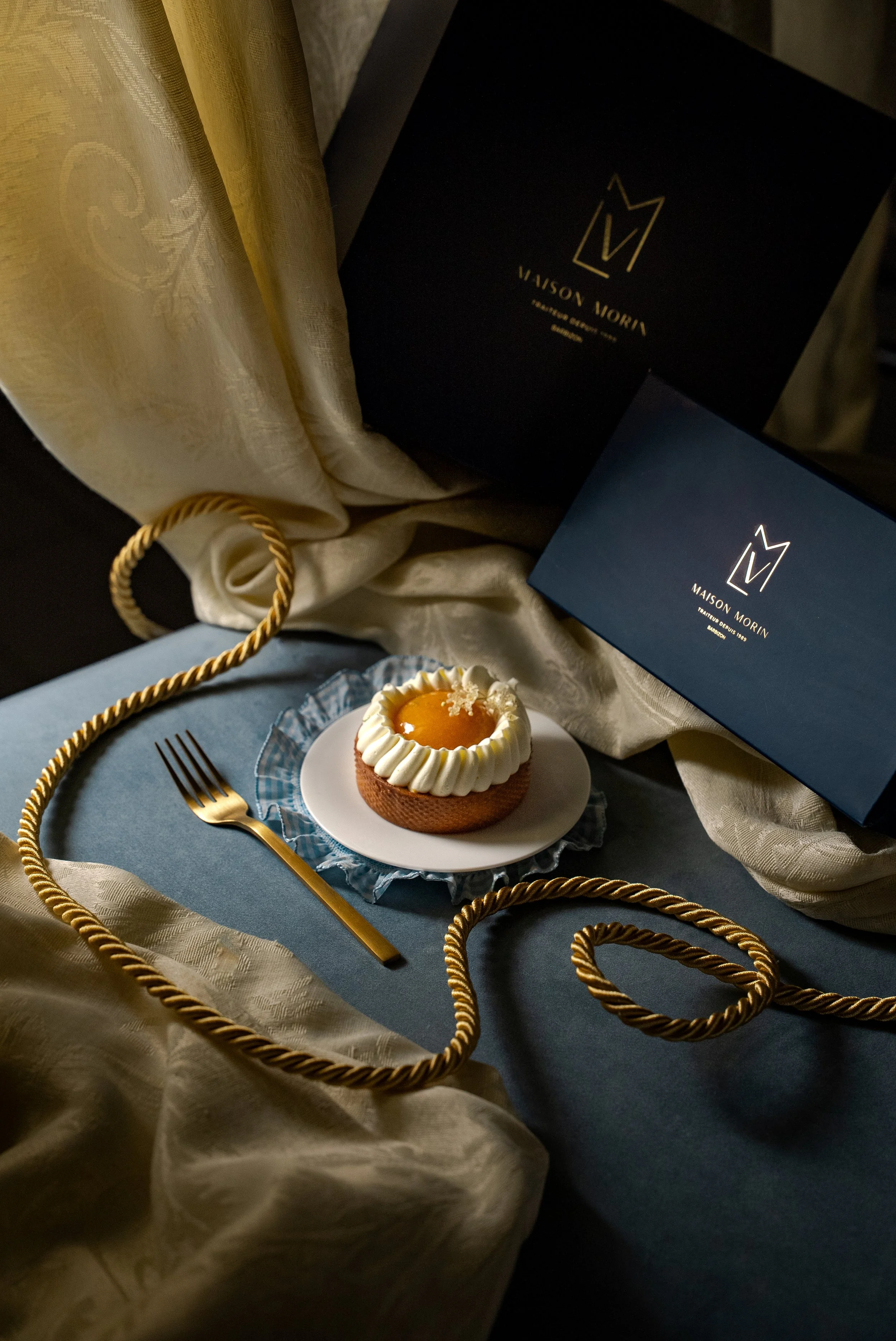 Un gâteau décoré avec de la crème et une confiture d'abricot, une fourchette dorée à côté, entouré de rubans dorés, avec des boîtes de Maison Morin en arrière-plan, le tout sur une surface en tissu bleu et des tissus dorés.