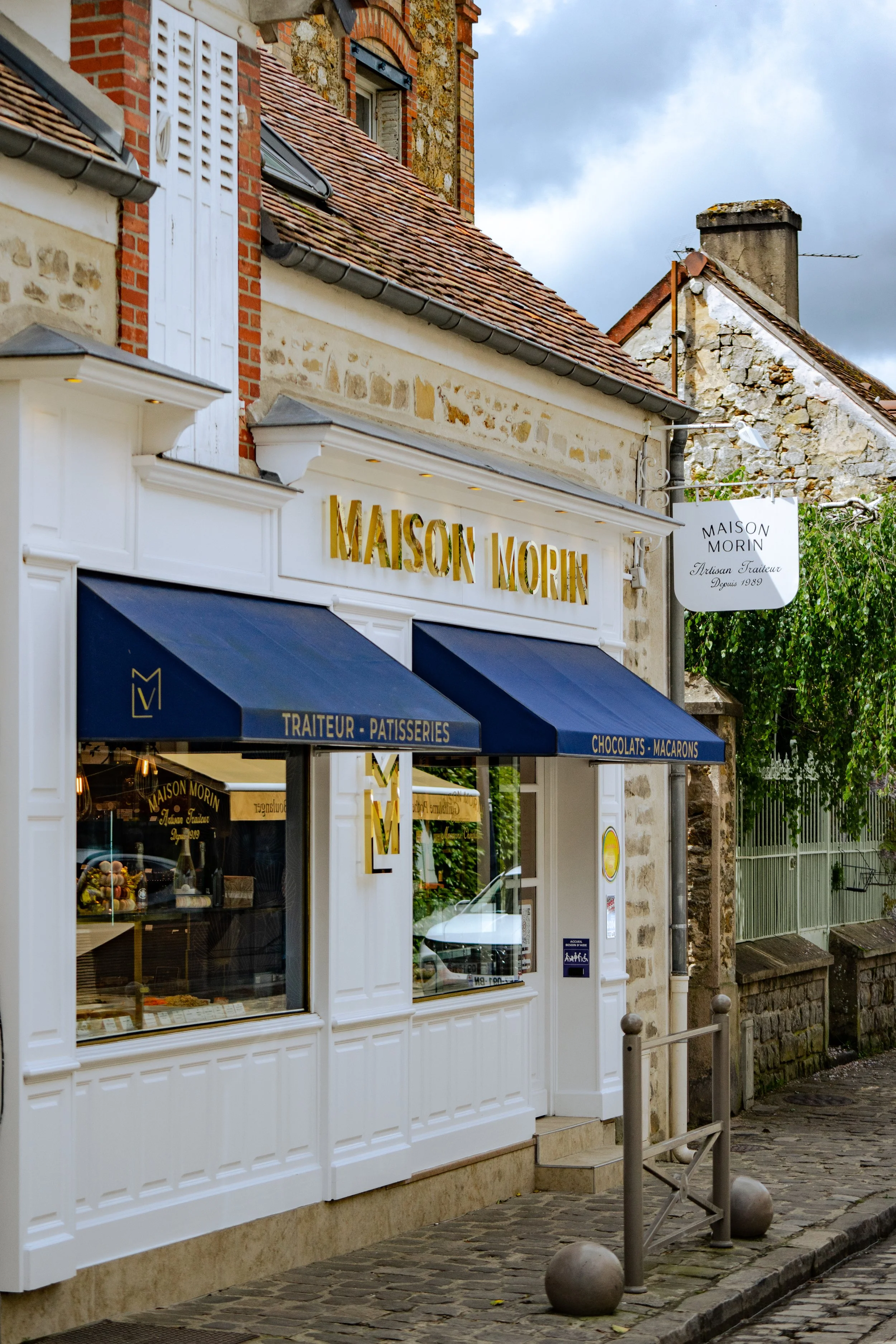 Vue de l'extérieur de la boutique Maison Morin avec enseignes en or, auvent bleu, fenêtres, bâtiment en pierre et rue pavée.