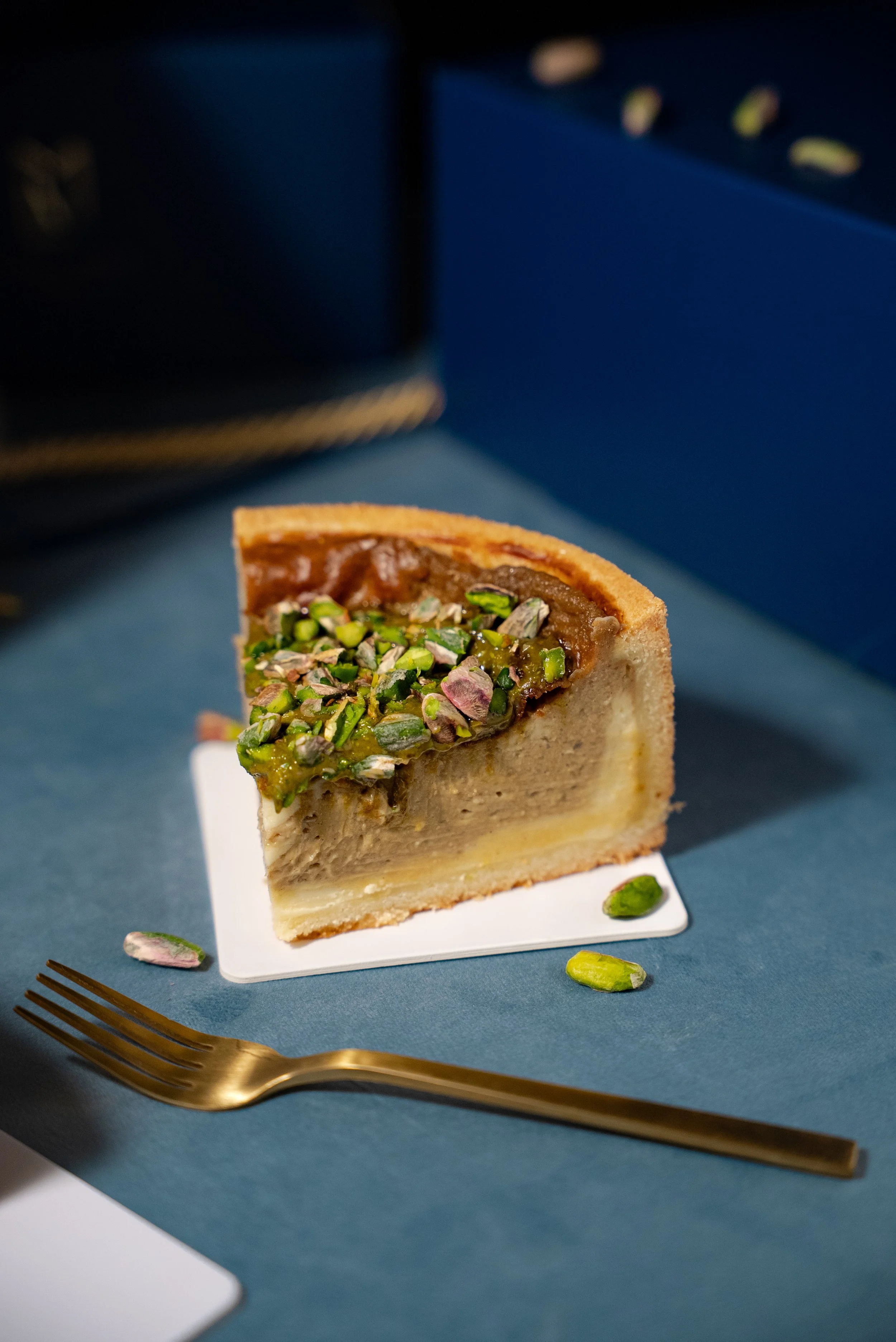 Part de tarte aux pistaches sur une assiette blanche, avec quelques pistaches autour, posée sur une nappe bleue, accompagnée d'une fourchette dorée.