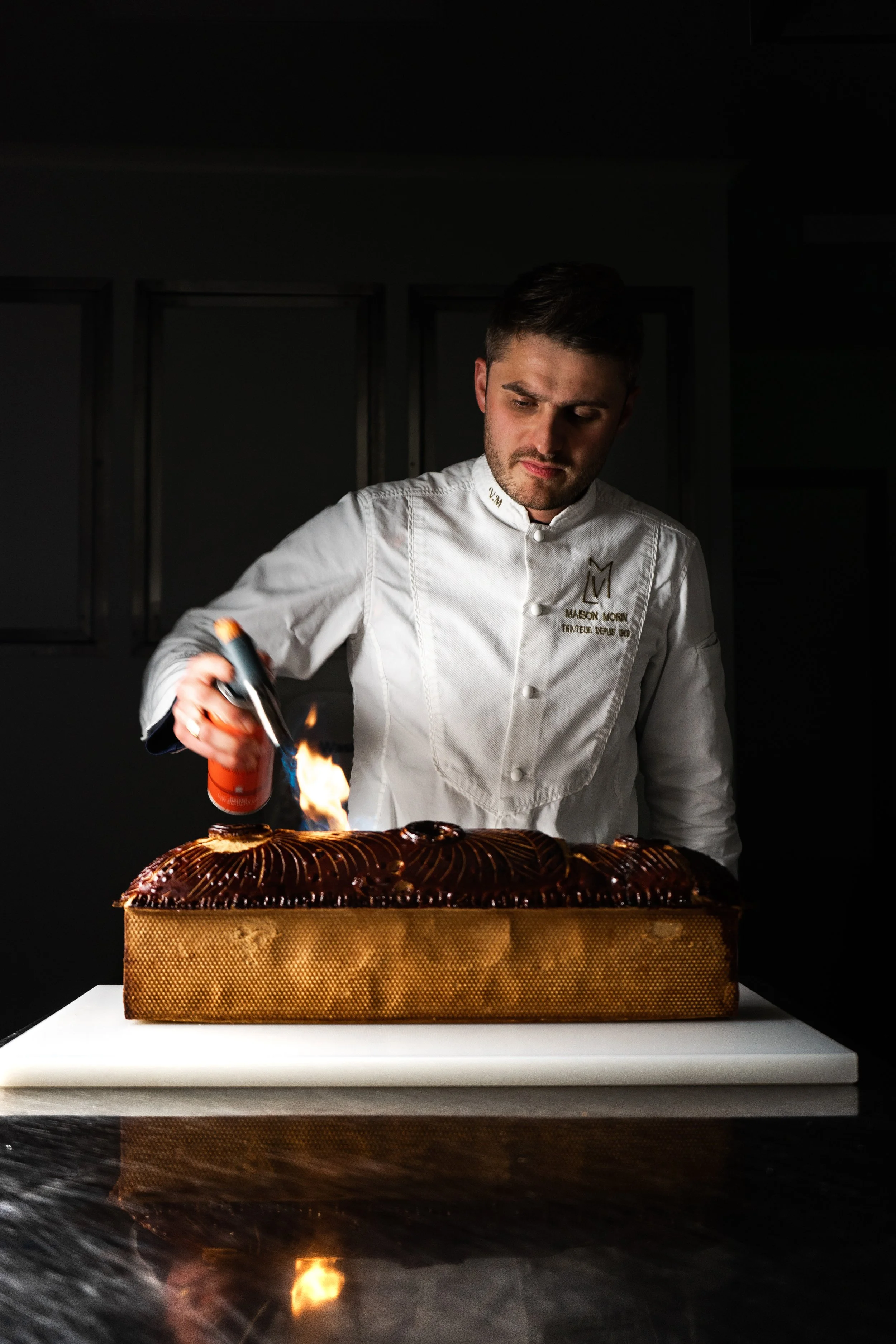 Un pâtissier, en robe blanche, utilisant un chalumeau pour flamber un gâteau décoré de glaçage chocolat, dans une cuisine sombre.