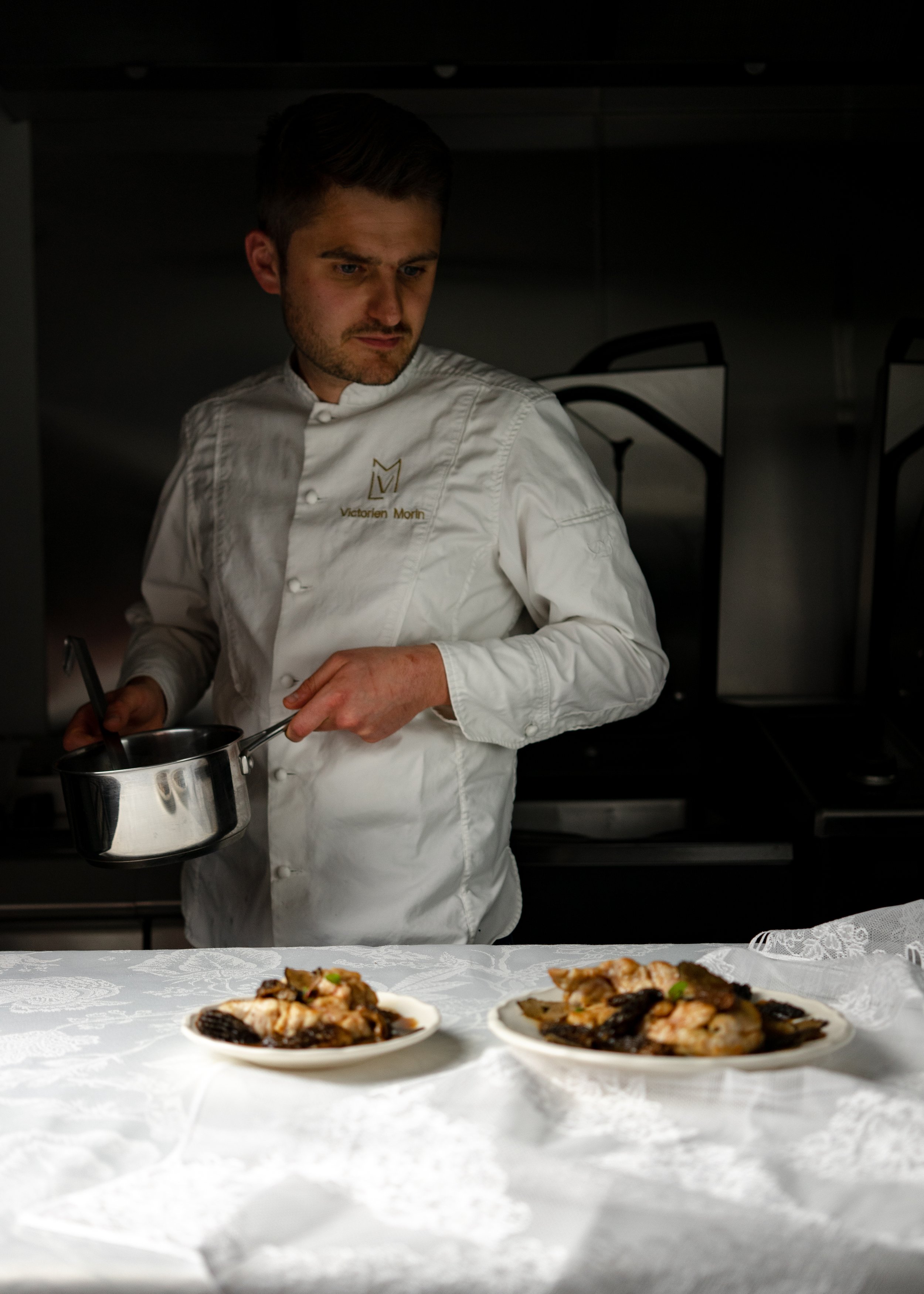 Chef en cuisine devant deux plats de nourriture sur une nappe blanche.