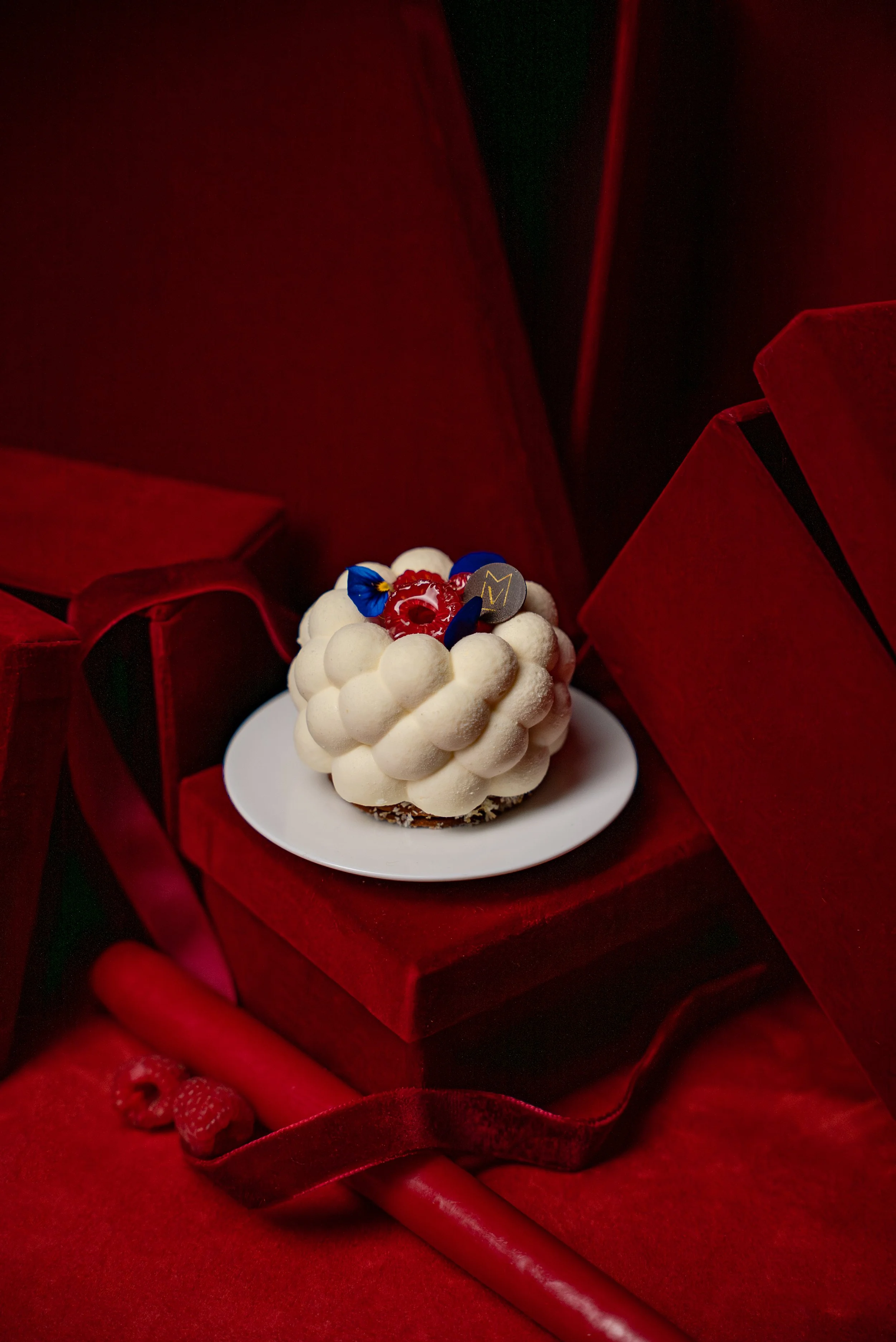 Gâteau blanc décoré avec des petits boules, garni de fruits rouges, de fleurs et de décoration en chocolat, présenté sur une assiette blanche, entouré de boîtes rouges en velours sur fond rouge.