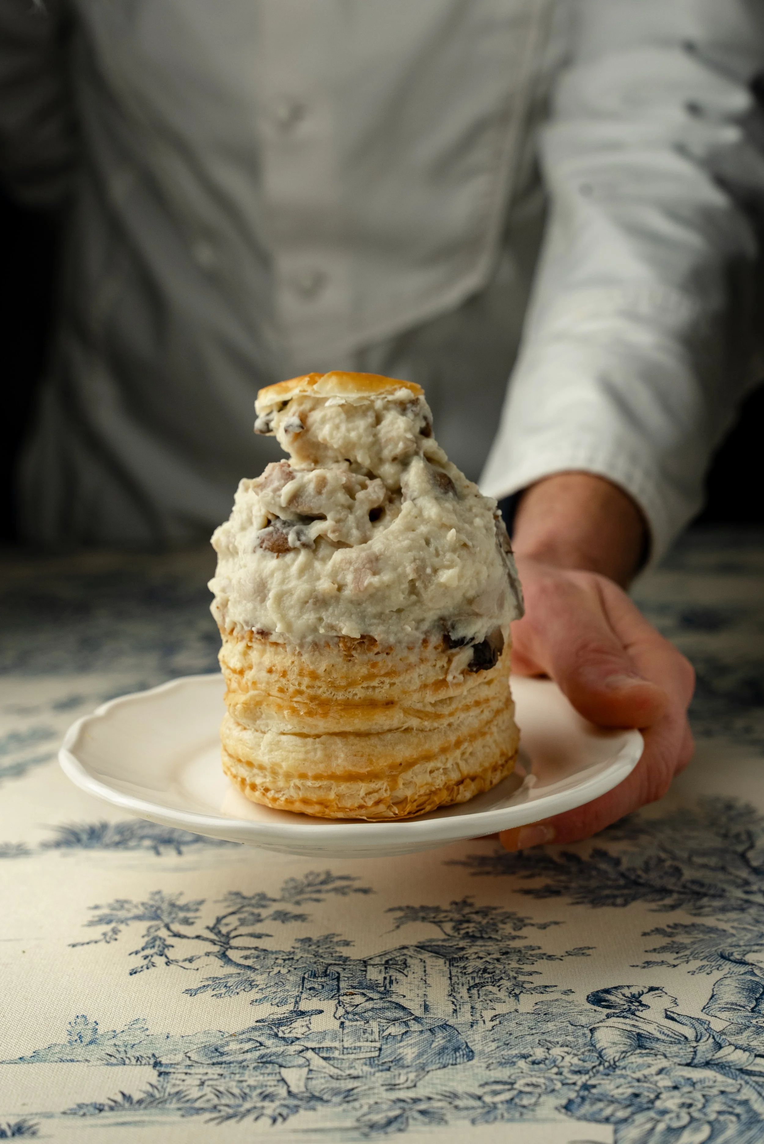 Person tenant une assiette avec un sundae de crème glacée aux morceaux de chocolat et biscuits, présenté sur une table à nappes avec motif paysage.