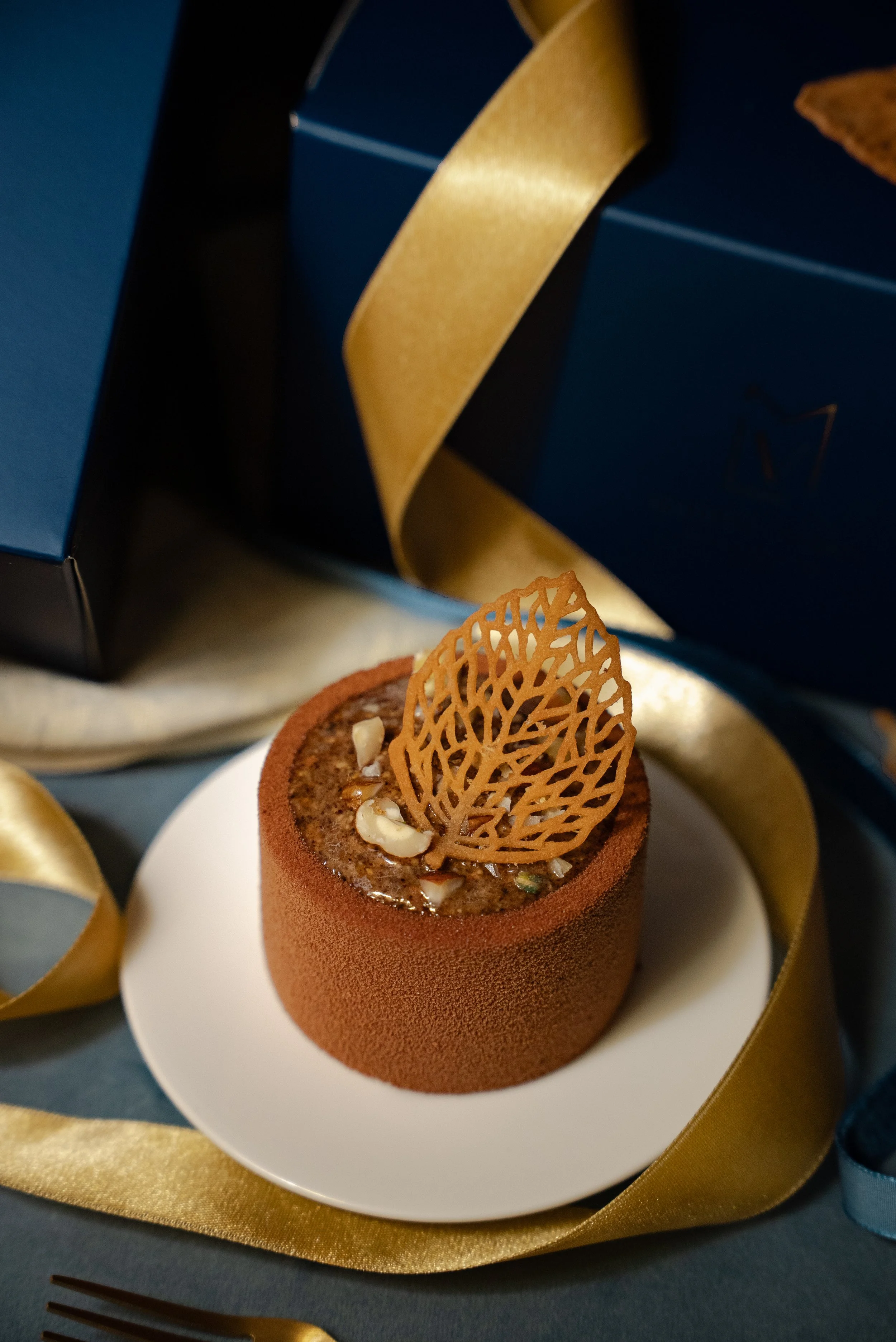 Gâteau au chocolat décoré d’une fine feuille en caramel, placé sur une assiette blanche, avec des boîtes en cadeau en arrière-plan, entouré de rubans dorés et bleus.
