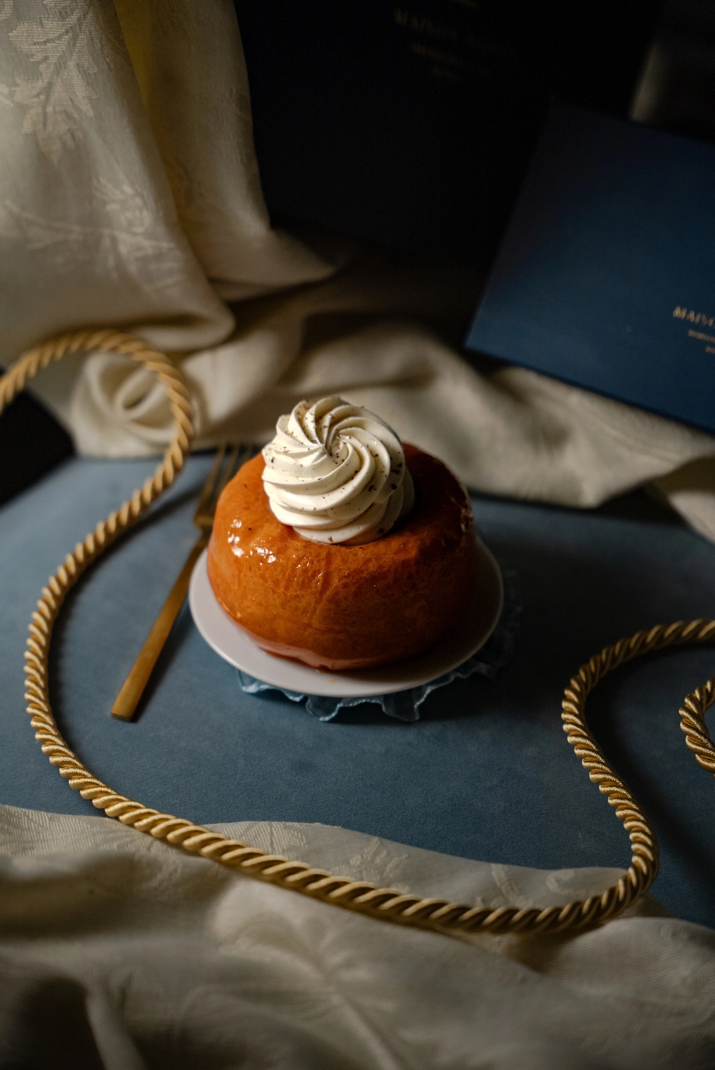 Gâteau rond avec de la crème fouettée en forme de spirale sur le dessus, posé sur une petite assiette blanche, entouré de ficelle dorée et posé sur une surface bleue, avec un drap beige en arrière-plan.