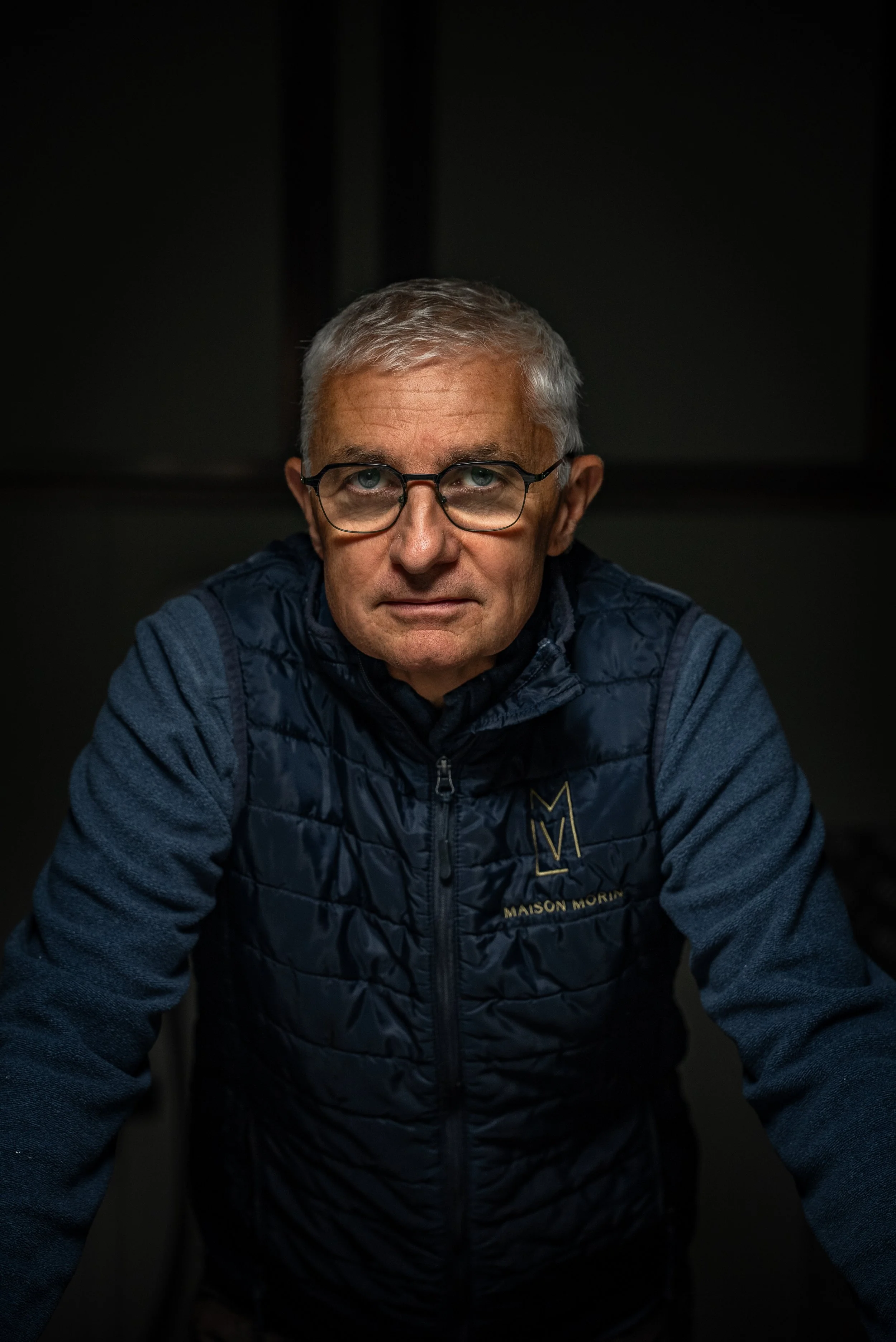Homme âgé portant des lunettes, vêtu d'une veste et d'un pull, face à la caméra dans un environnement sombre.