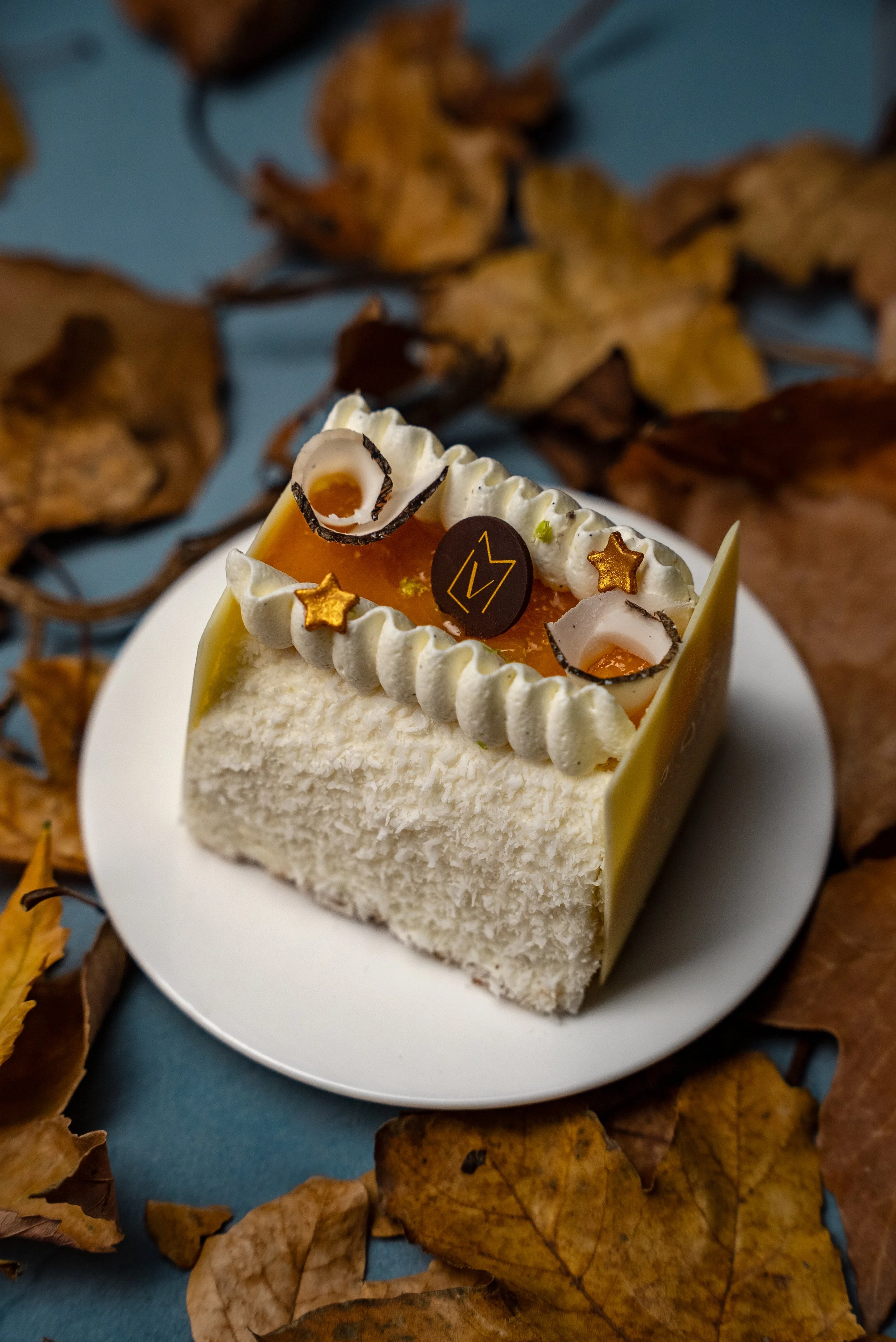 Gâteau avec une inclination de style anniversaire, doté de crème blanche, d’une couche de gelée d’orange, ornementé de décoration en chocolat en forme d’étoile et de boucle, et entouré de feuilles d'automne sèches sur fond bleu.