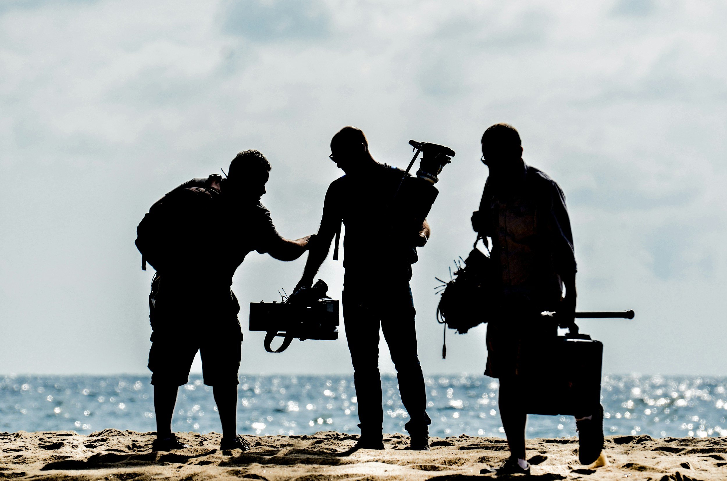 Silhouettes of three filmmakers on a beach, preparing equipment with the ocean in the background.