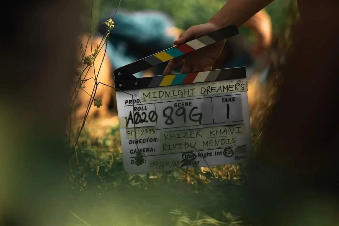 Clapperboard for the film "Midnight Dreamers" held by a person outdoors, surrounded by grass and plants.
