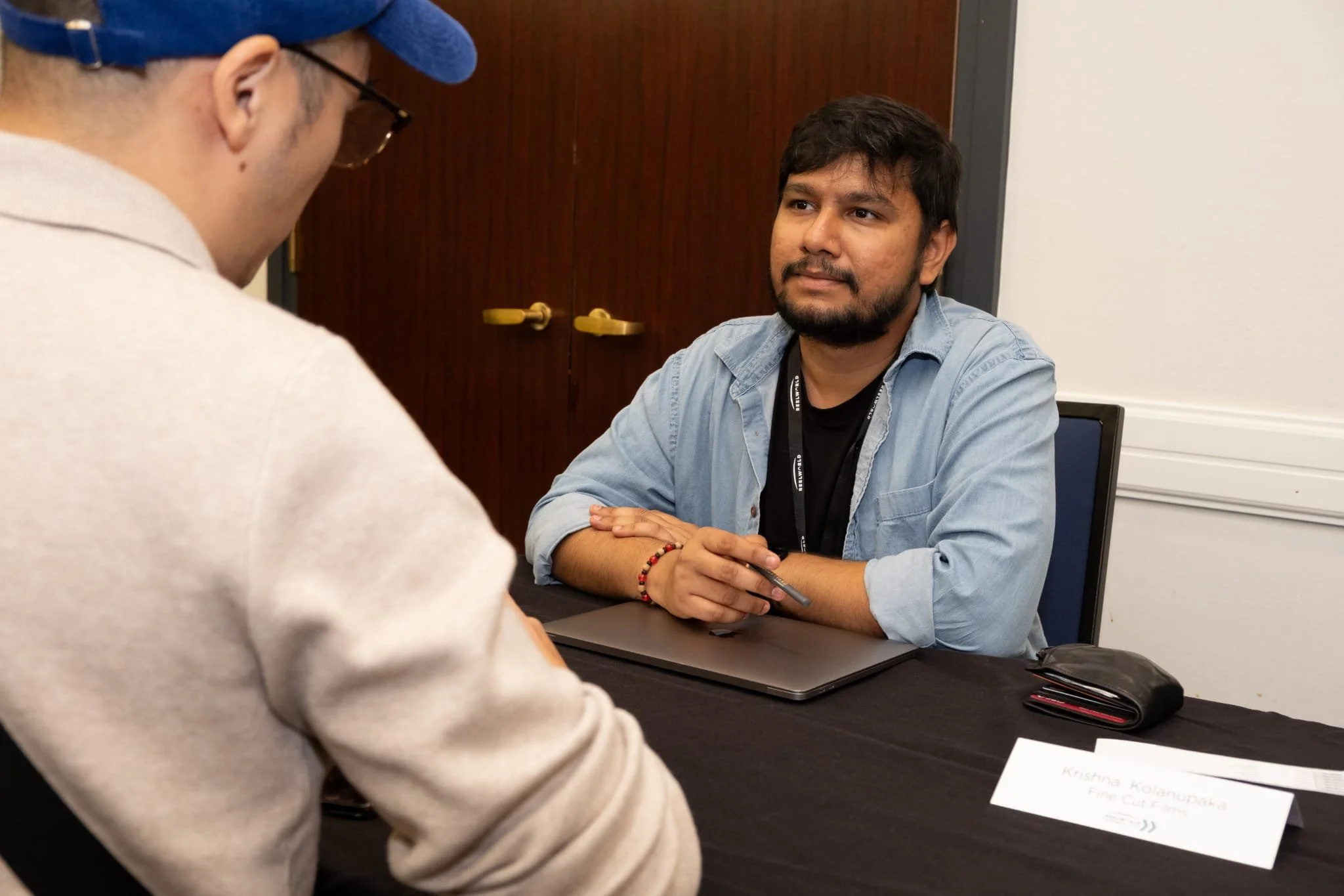 An interview or meeting taking place with Krishna sitting at a table and speaking to a person wearing glasses and a hat.