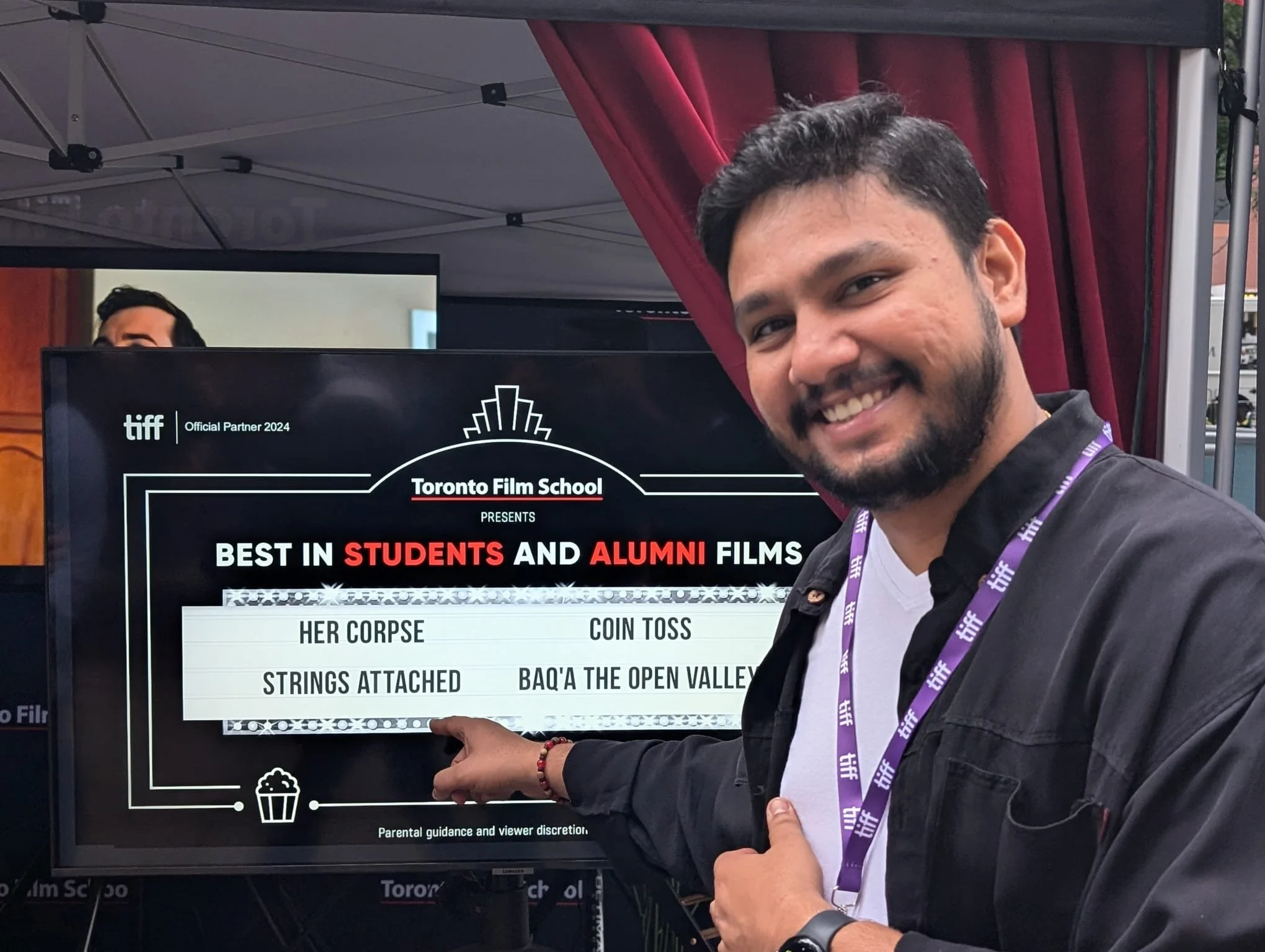 Krishna is smiling and pointing at a screen displaying award-winning student and alumni films at the Toronto Film School. The display lists four titles: Her Corpse, Coin Toss, Strings Attached, and Baq'a the Open Valley.