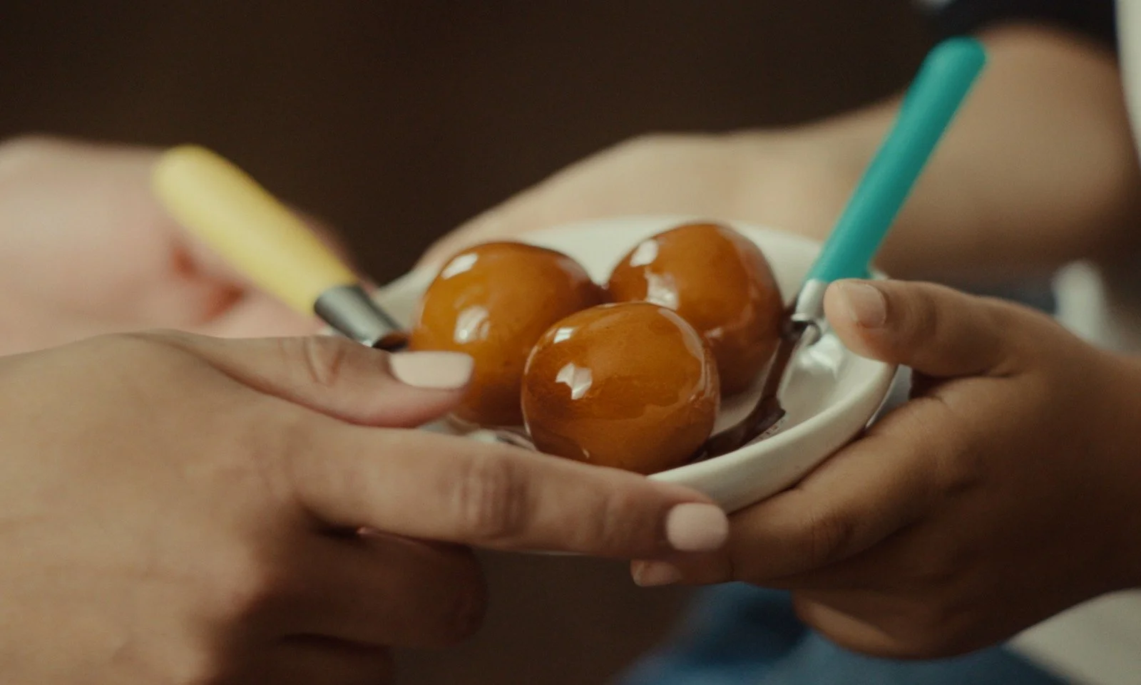 Close-up of a person's hand holding a small white dish with four glossy, round, caramel-colored candies and two tongs, one yellow and one teal.