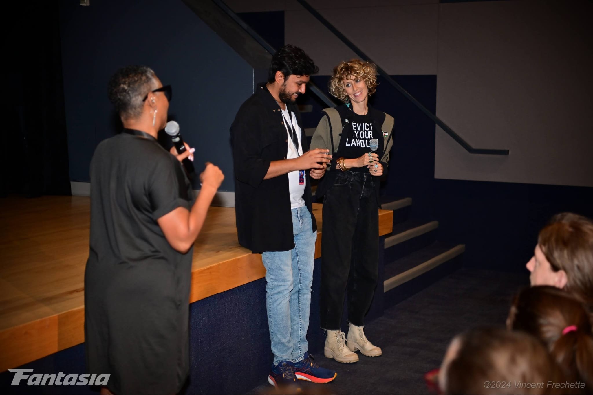 Two women and Krishna standing on stage with microphones; one woman speaking to an audience, the other listening; audience members in foreground