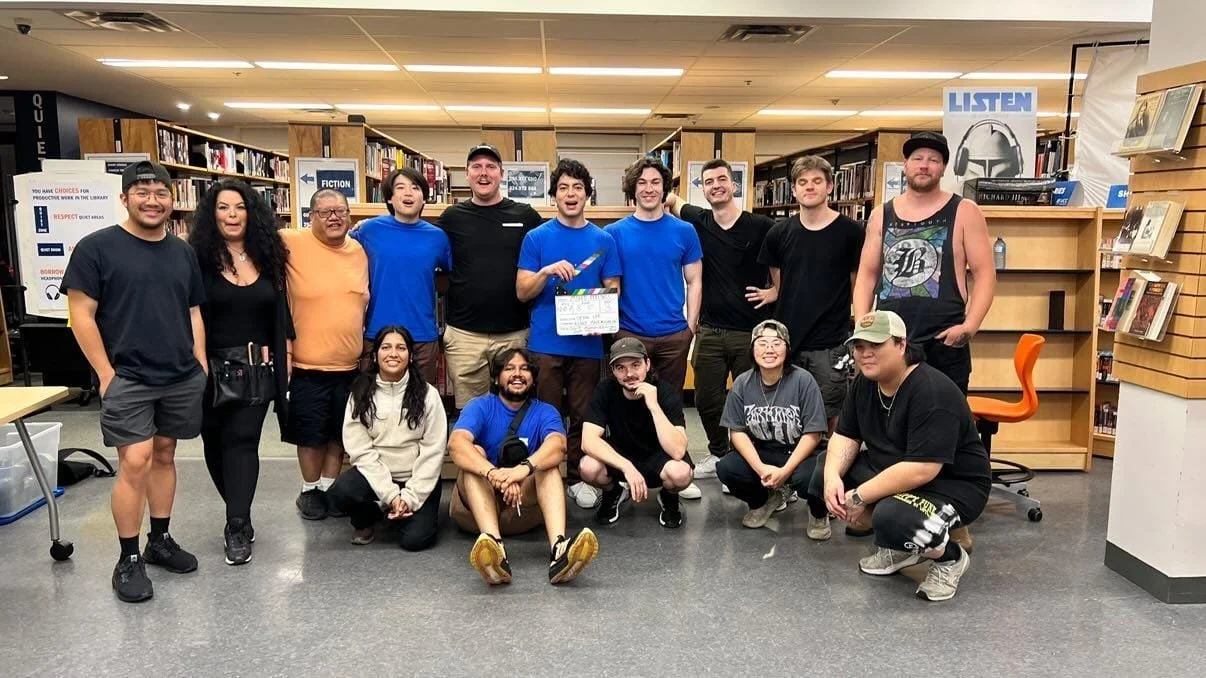 Group of 14 people including Krishna posing for a photo inside a library, some standing and some squatting, with bookshelves and signs in the background.