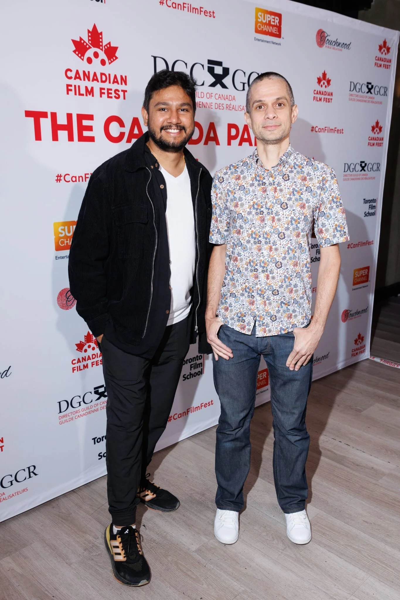Krishna and his friend standing in front of a banner promoting the Canadian Film Fest, smiling at the camera. One man is wearing a black jacket, white T-shirt, and sneakers, while the other is wearing a floral shirt, jeans, and white shoes.