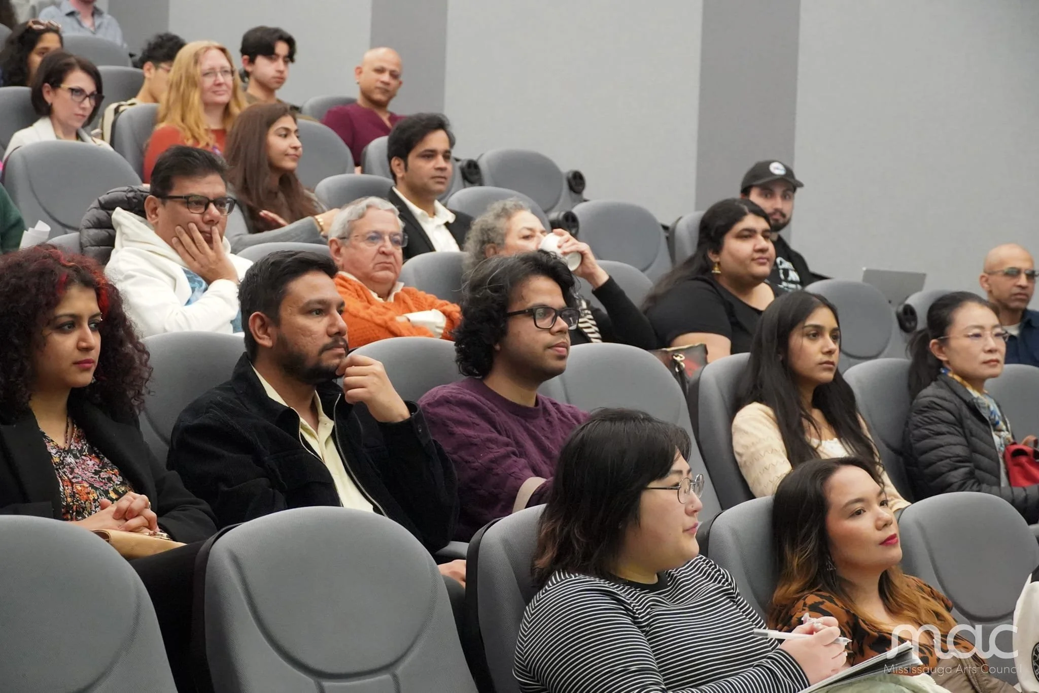 An audience including Krishna and his frequent collaborator Anubhav Singh seated in a conference room, listening attentively to a presentation. The room has gray walls and gray chairs arranged in rows.