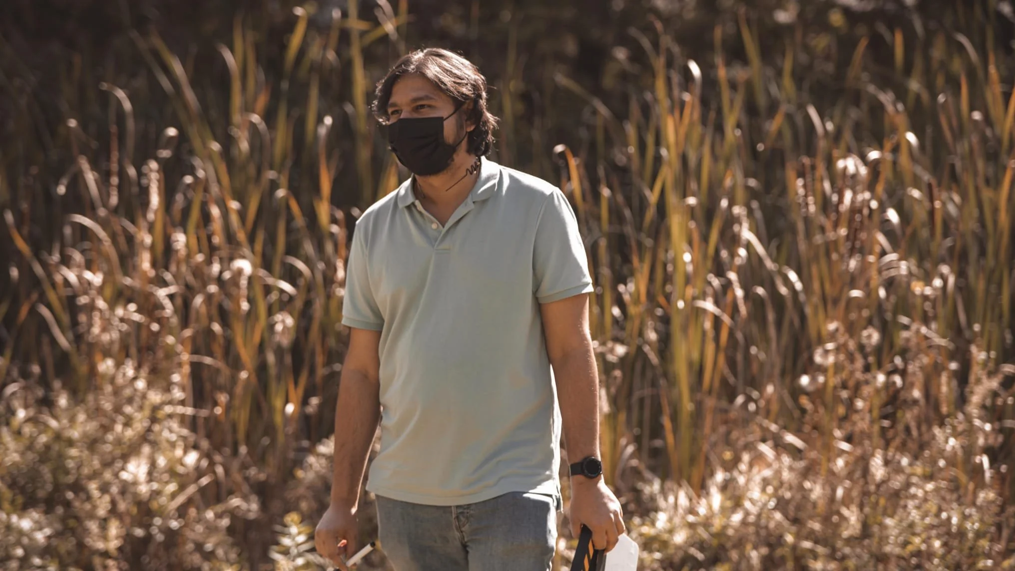 Krishna wearing a black face mask and a light-colored polo shirt walking through a field of tall, dry grass or crops, holding a small object or device in his right hand and a white paper or card in his left hand.