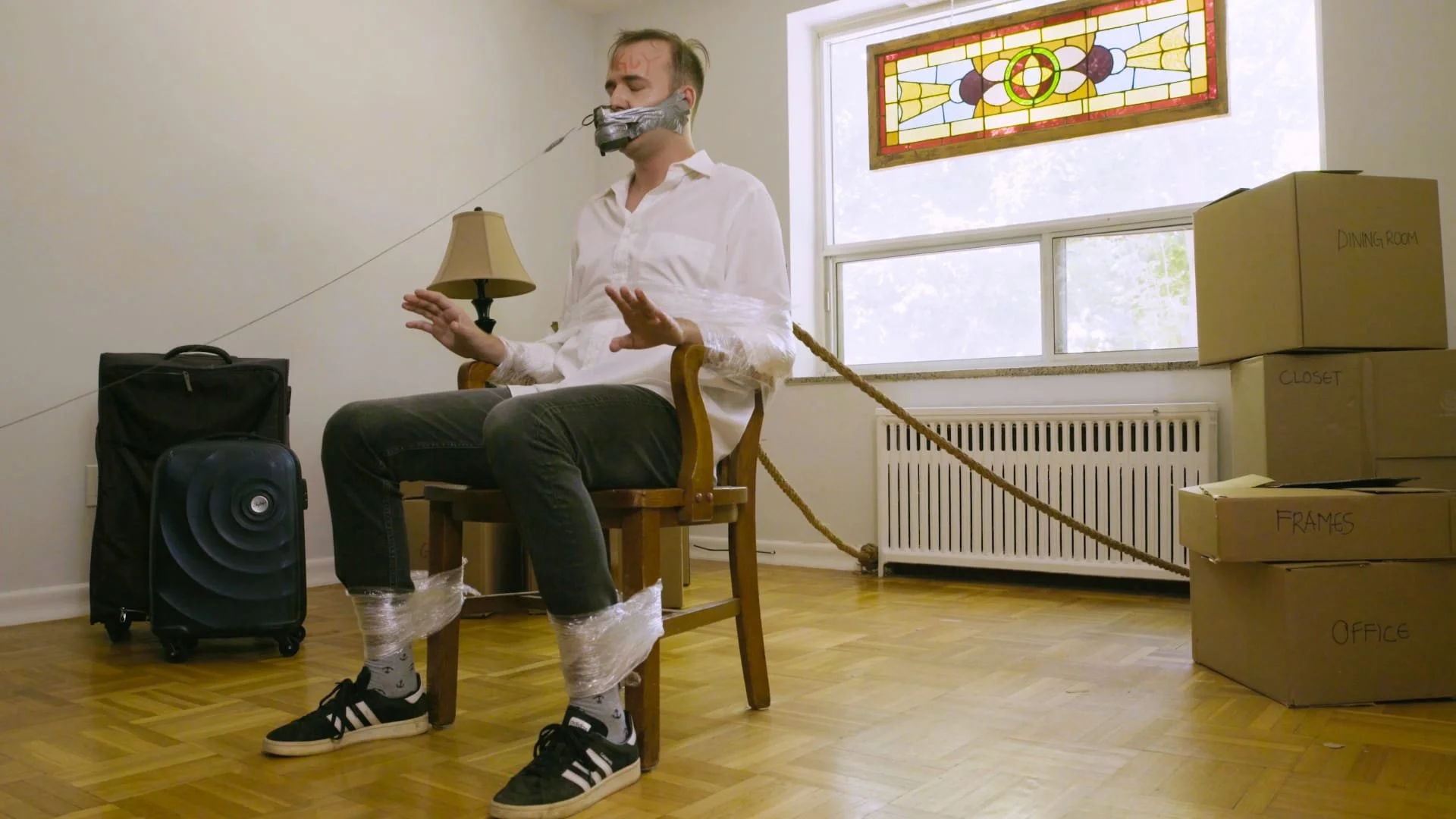 Man tied to chair with rope, wearing tape over mouth, in a room with boxes labeled 'dining room,' 'closet,' 'frames,' and 'office,' and a stained glass window.