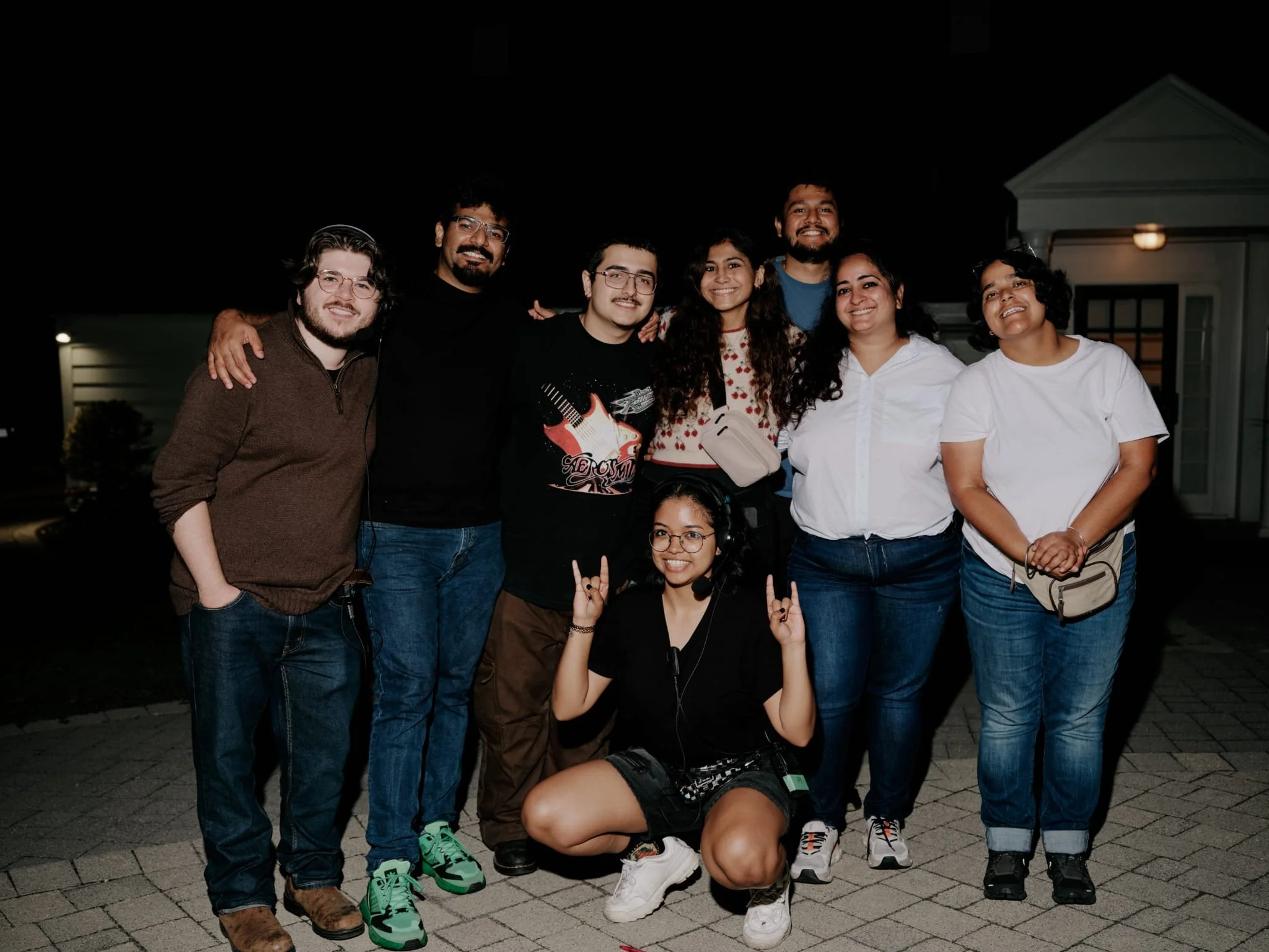 Group of eight diverse friends smiling and posing together outdoors at night, with some wearing casual clothing and one person in the front sitting on the ground making rock hand gestures.