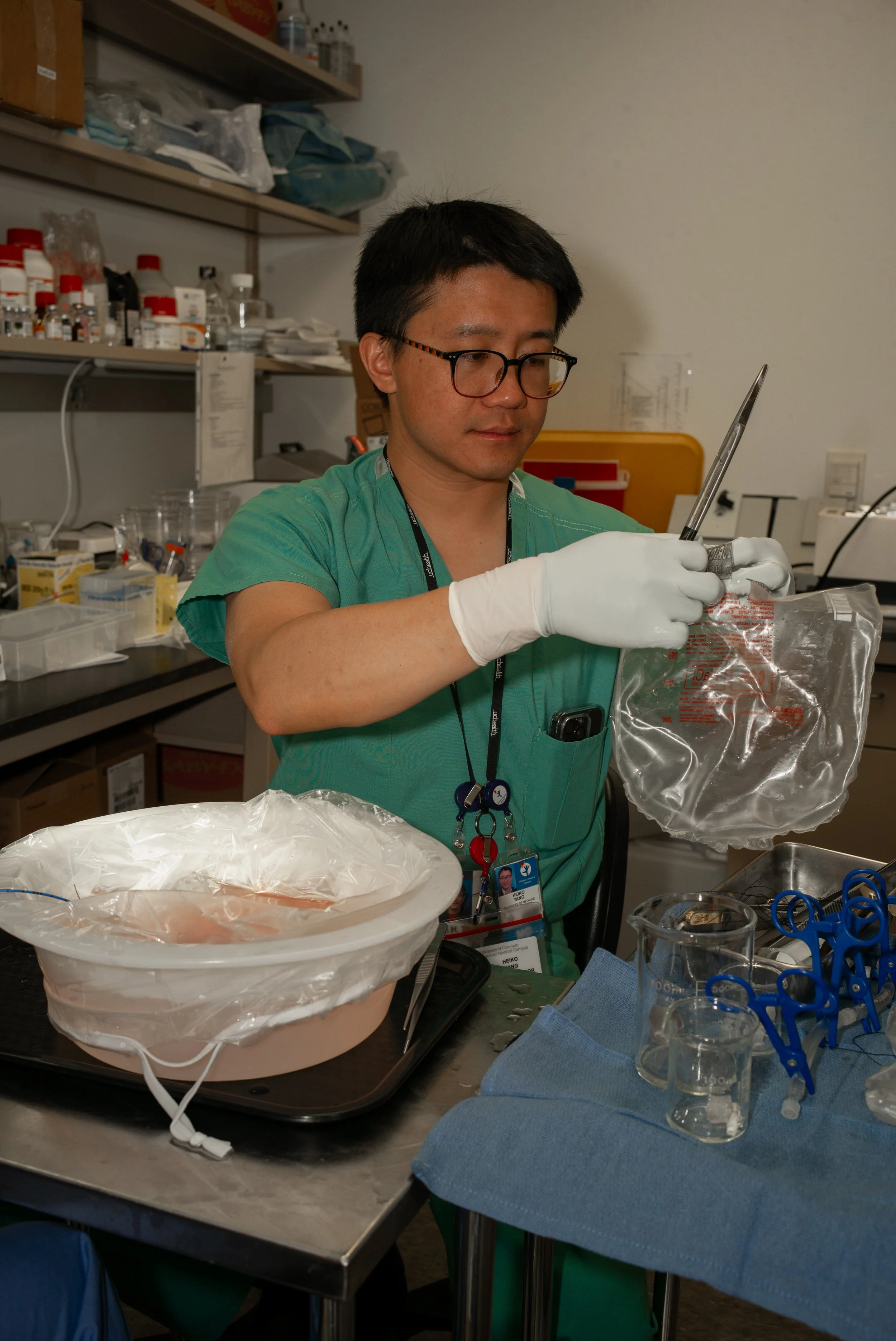 A scientist in green scrubs and white gloves working in a laboratory, handling a sealed plastic bag with medical or scientific supplies, surrounded by beakers, scissors, and lab equipment.