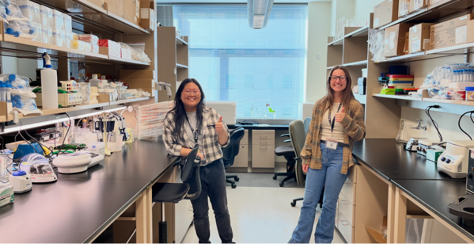 Two women smiling and giving thumbs-up in a laboratory with shelves filled with scientific supplies and equipment.