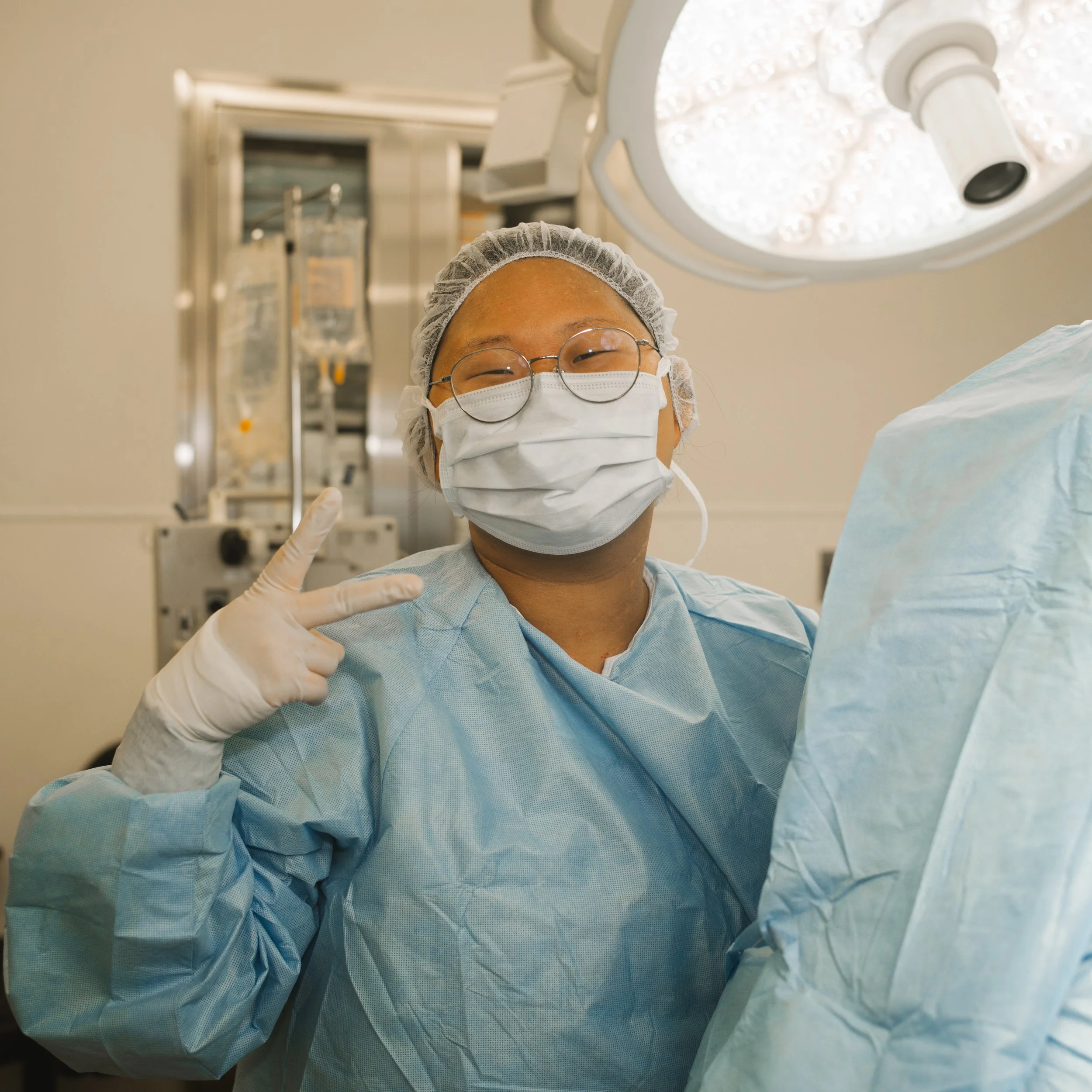 An Asian medical professional wearing scrubs, a face mask, glasses, and a hair cap, smiling and making a peace sign in an operating room with surgical lights overhead.