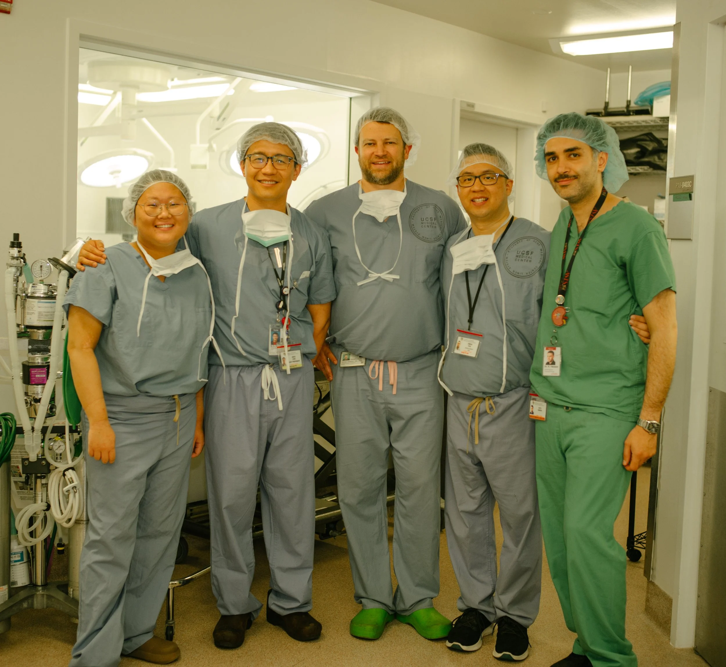 Group of five medical professionals, four wearing gray surgical scrubs and one in green scrubs, standing together in a hospital operating room, smiling.