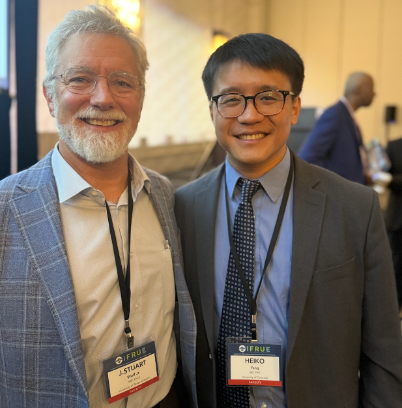 Two men in business suits smiling at a conference, wearing name badges.