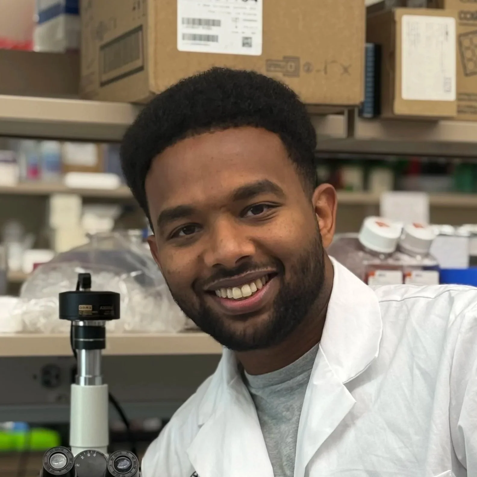 Young man smiling in a laboratory, wearing a white lab coat, with scientific equipment and supplies in the background.