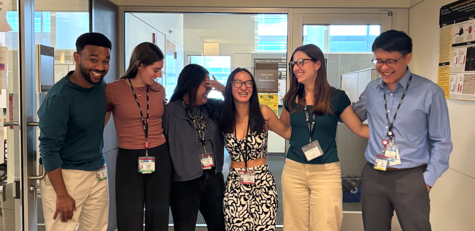 Six diverse people in professional attire standing together in an office or conference setting, smiling and laughing.