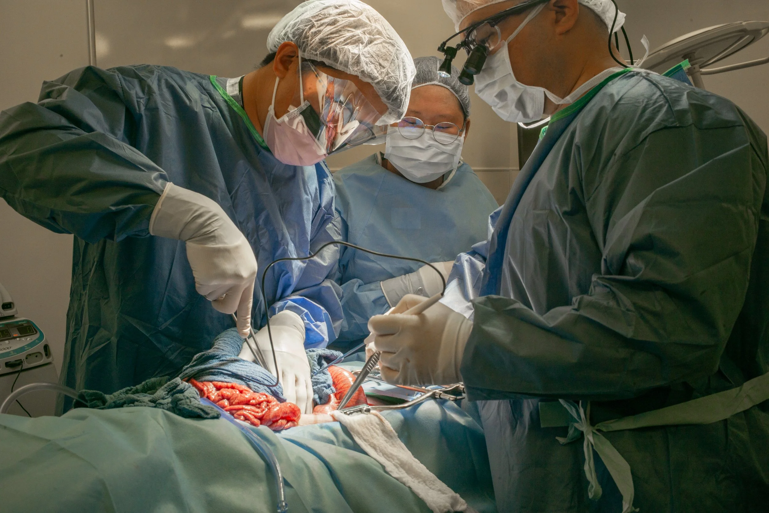 Medical team performing surgery in an operating room, surgeons wearing scrubs, masks, gloves, and surgical headlamps, focused on a patient's abdomen.