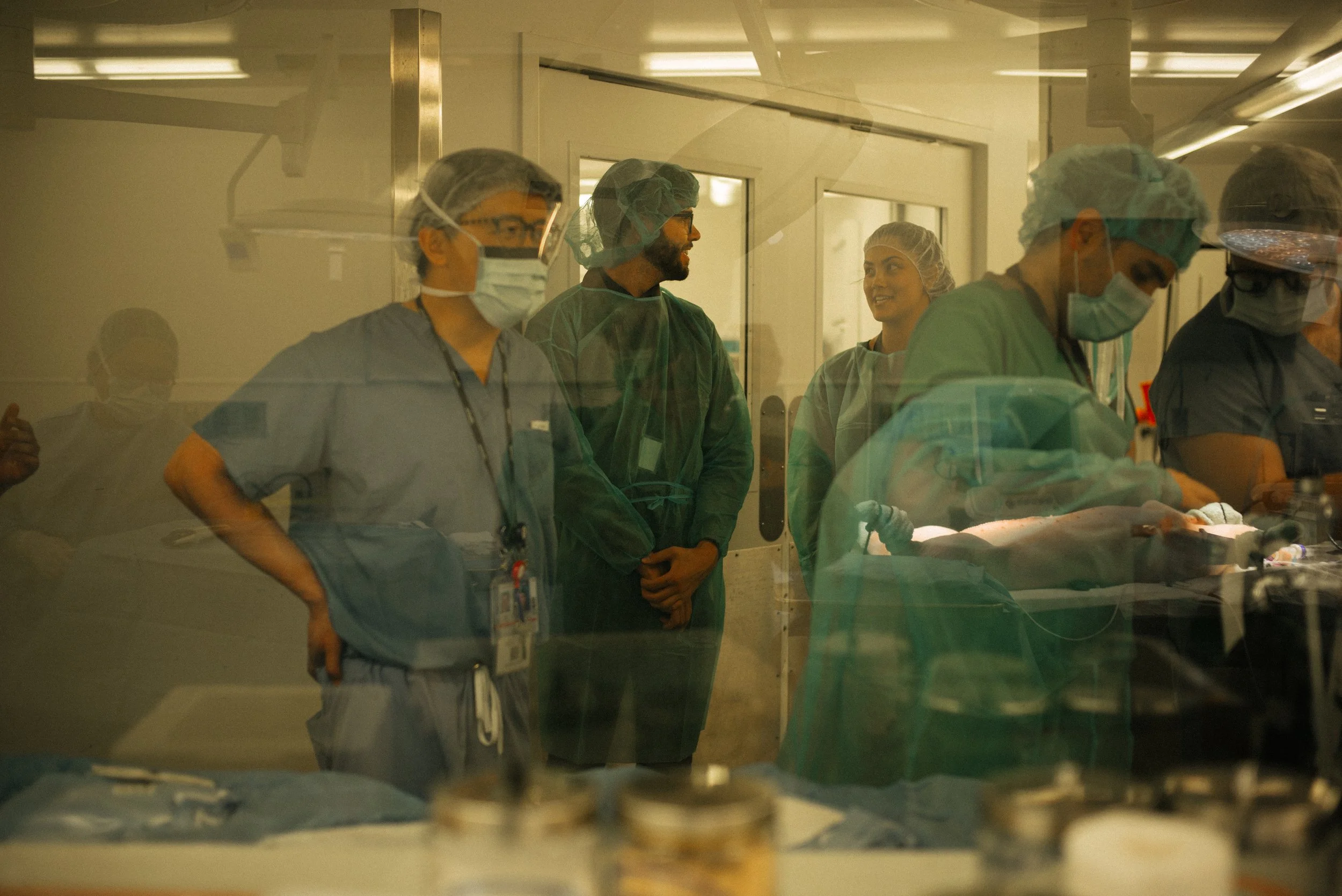 Medical professionals in scrubs and masks working in an operating room, viewed through a glass window.