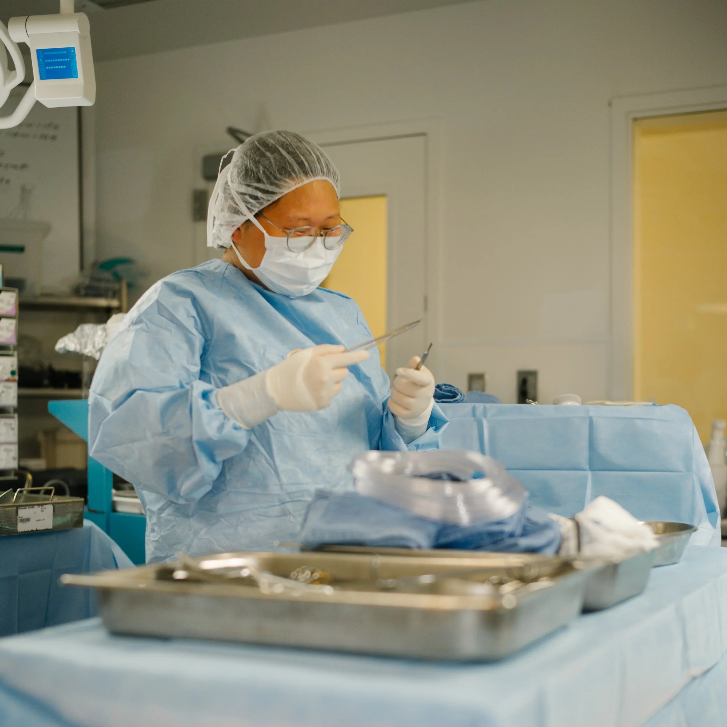 A medical professional wearing scrubs, gloves, a mask, hairnet, and glasses stands at a surgical table in a hospital, preparing tools for a procedure.