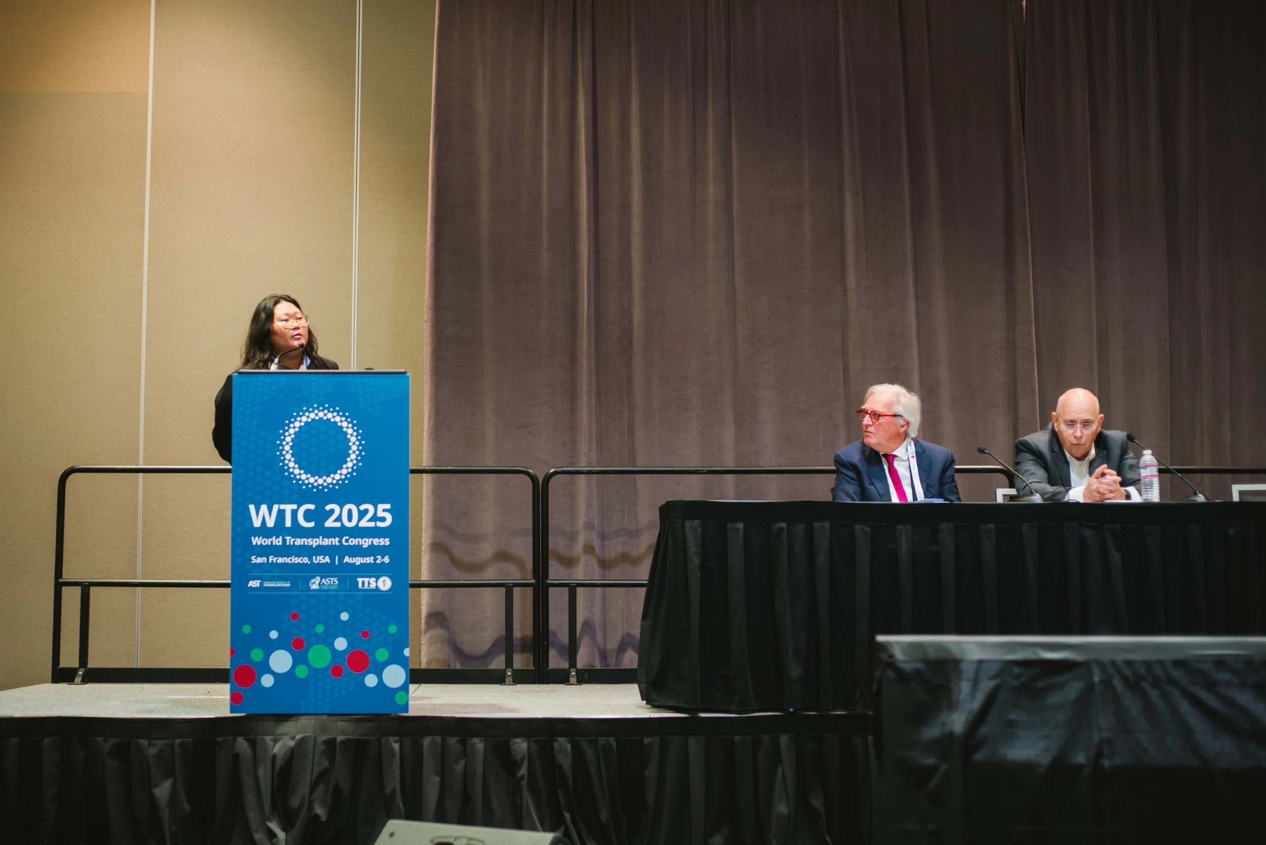 A woman standing at a podium presenting at the WTC 2025 World Transplant Congress in San Francisco, USA, August 2-6, with two men seated at a table listening.