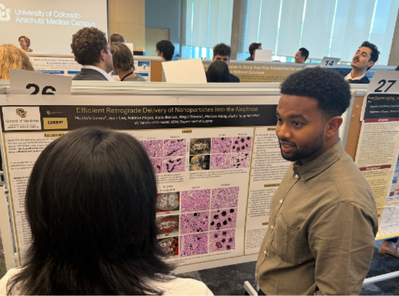 Two people discussing a science research poster titled 'Efficient Detergent-Enabled Delivery of Nanoparticles into the Alveolar' at a scientific conference at the University of Colorado Anschutz Medical Campus, with other attendees and posters in the background.