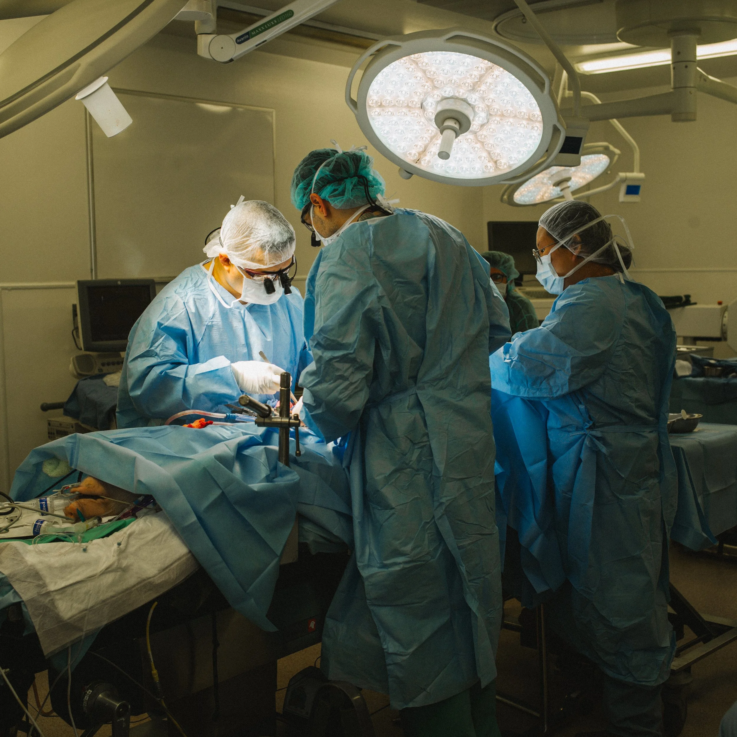 A team of surgeons performing an operation in an operating room, all wearing surgical gowns, masks, and protective eyewear, under bright overhead surgical lights.