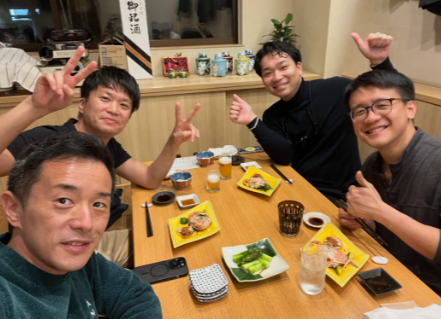 Four men sitting at a restaurant table with plates of food, drinks, and chopsticks, smiling and making peace signs.