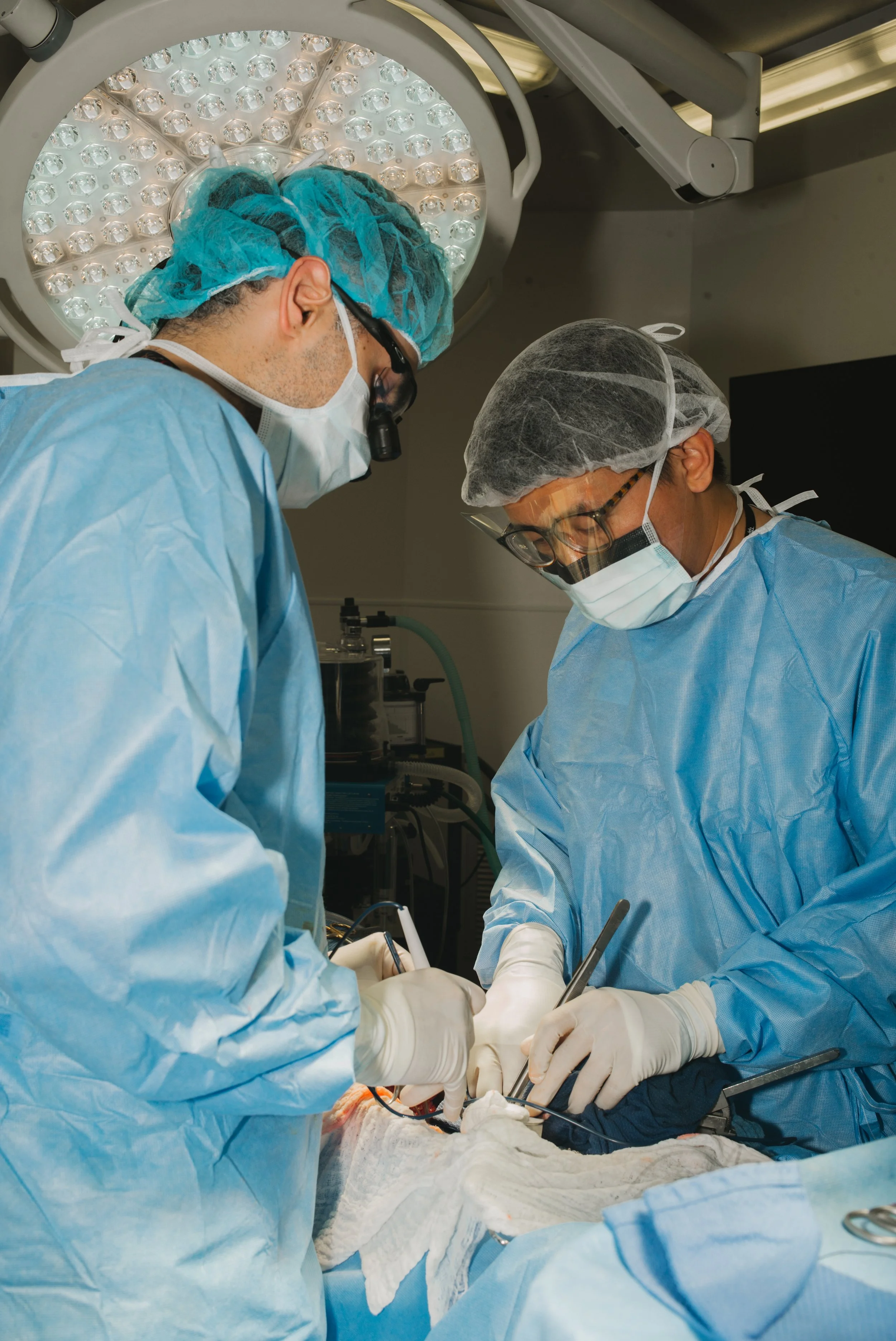 Two surgeons in blue scrubs and masks performing surgery in an operating room, with bright surgical lights overhead and medical equipment in the background.