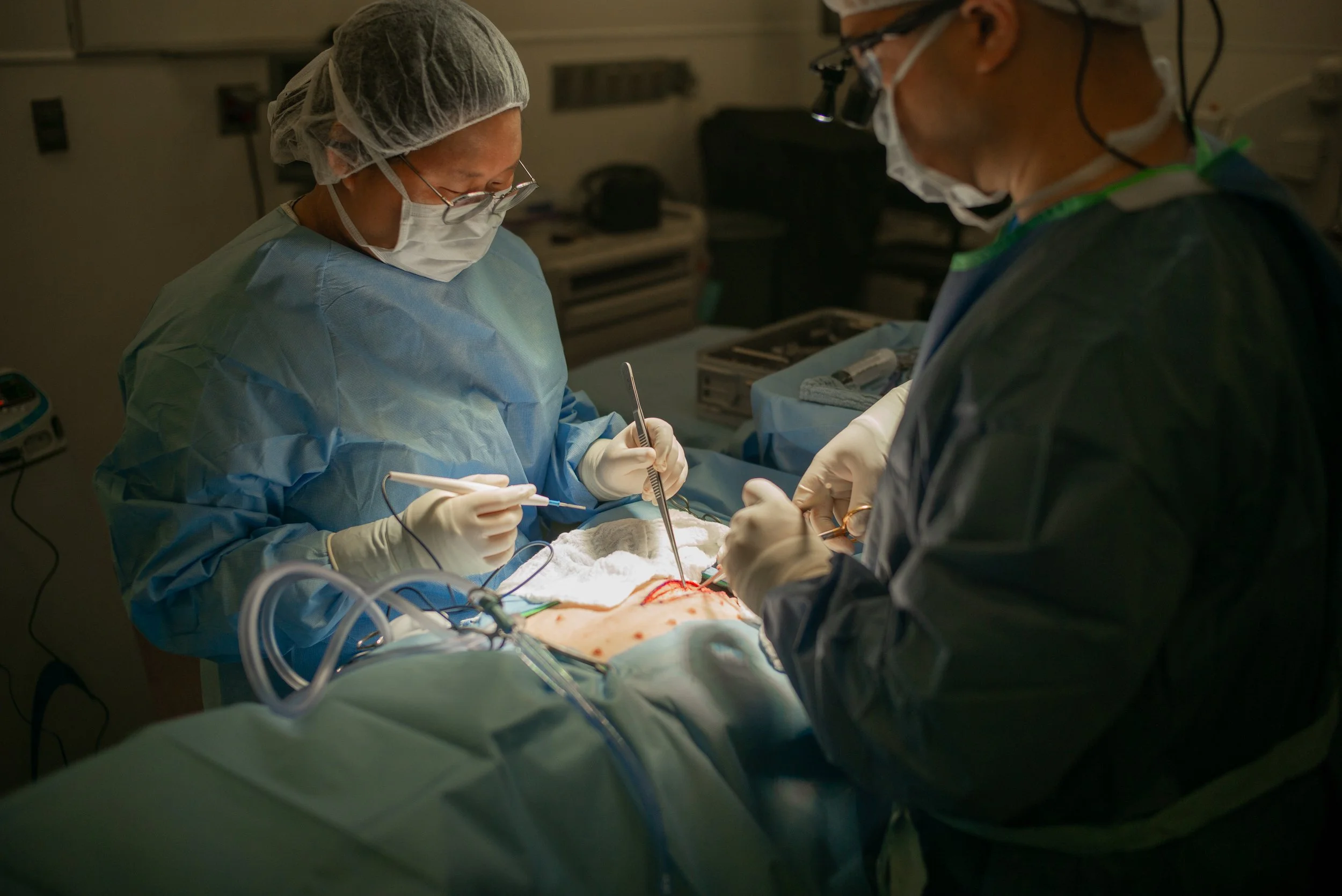 Two surgeons performing surgery in an operating room, dressed in sterile blue and green surgical gowns, masks, and gloves, focused on the procedure.