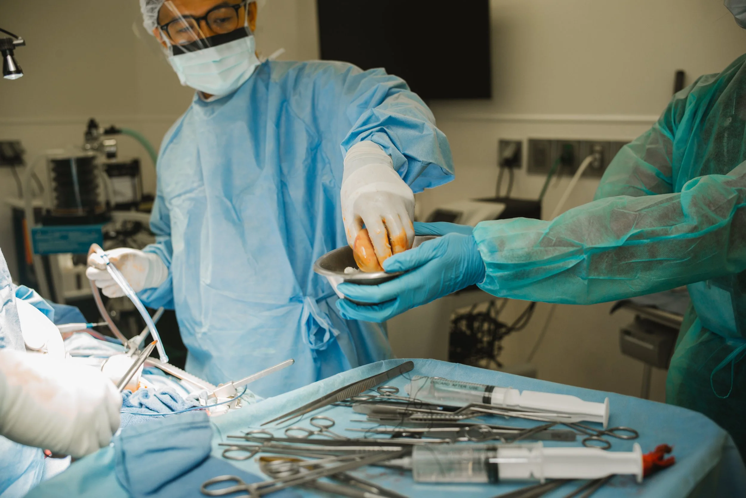 Surgeons in blue scrubs, gloves, masks, and protective eyewear performing a surgical procedure in an operating room with medical instruments and equipment on a sterile table.