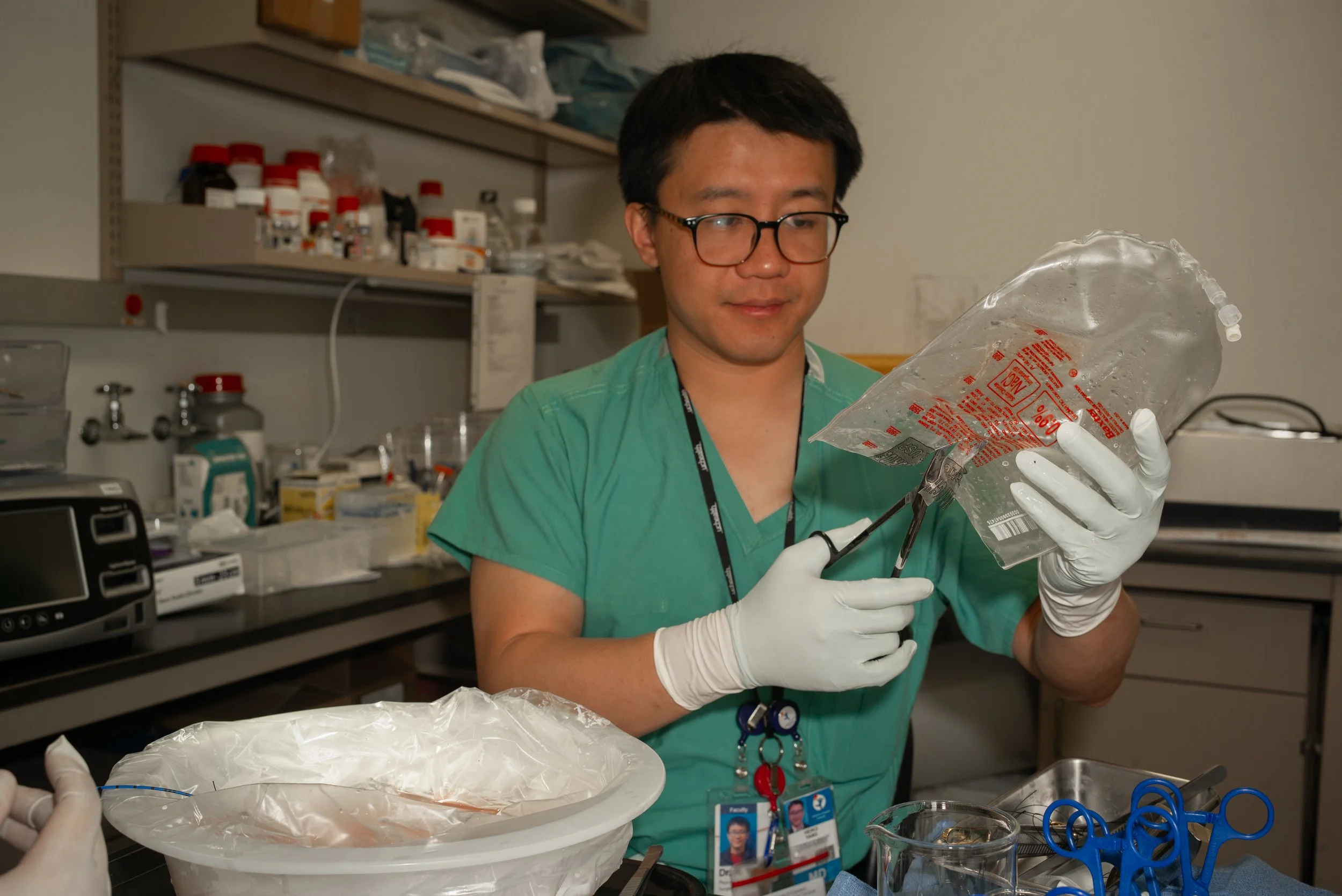 Medical professional wearing glasses, green scrubs, and white gloves, preparing medical supplies in a clinical setting.