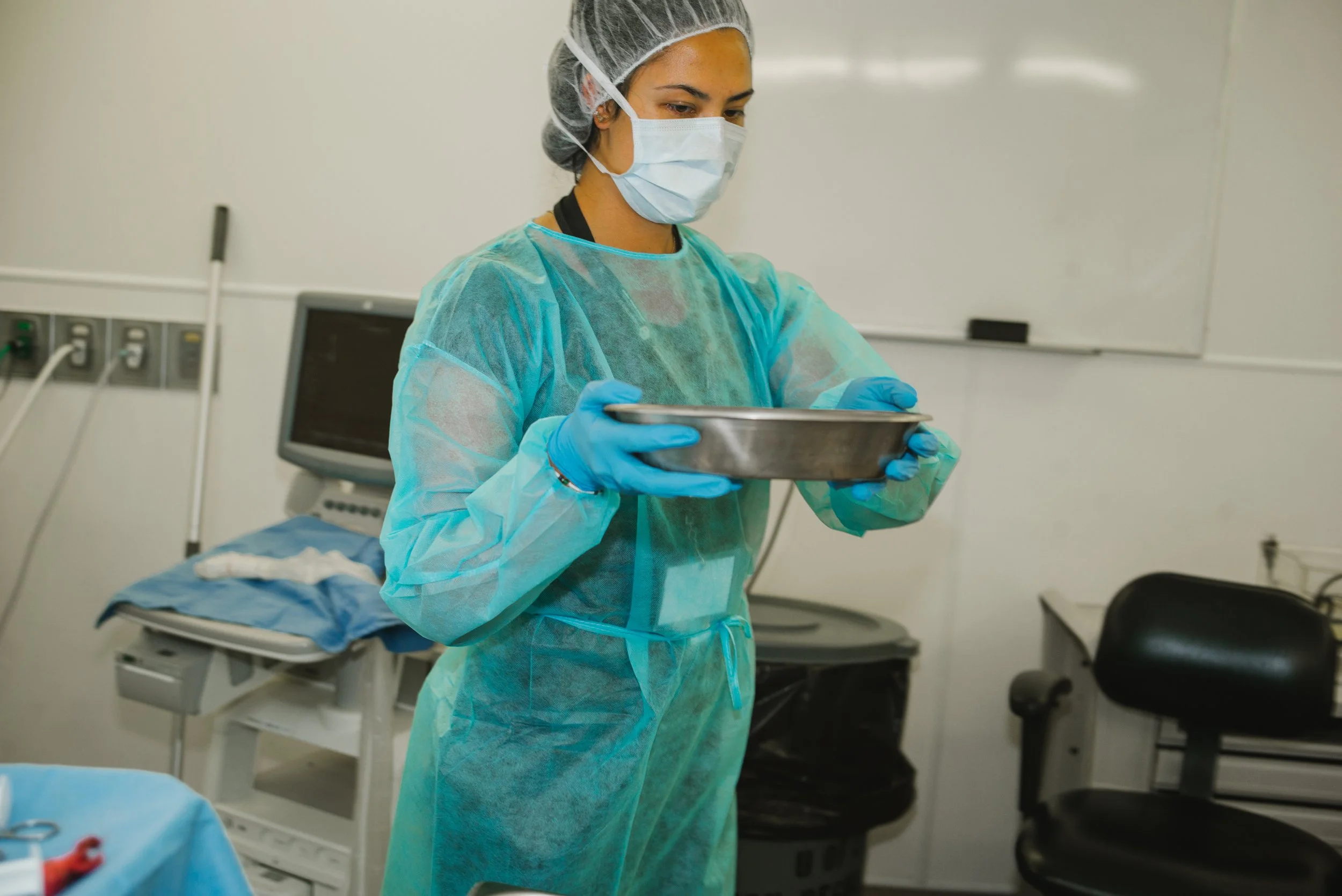 A female healthcare worker wearing a mask, gloves, and protective gown, standing in a clinical setting, examining a silver tray.