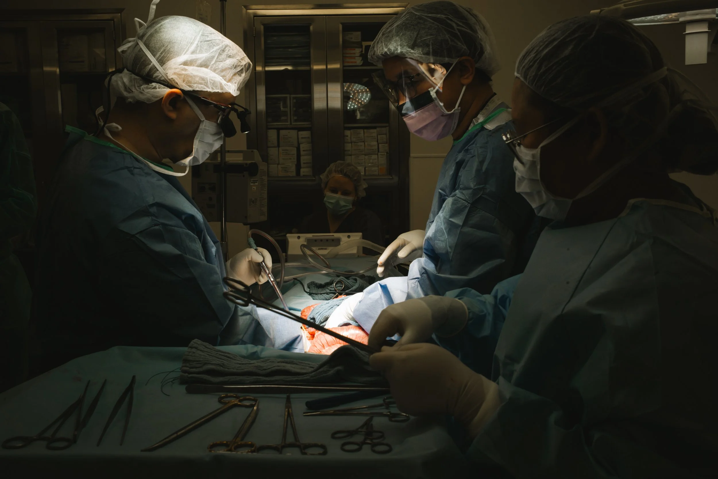 A team of healthcare professionals, dressed in surgical scrubs, masks, and gloves, performing surgery in an operating room. Surgical instruments are laid out on a sterile table in the foreground.