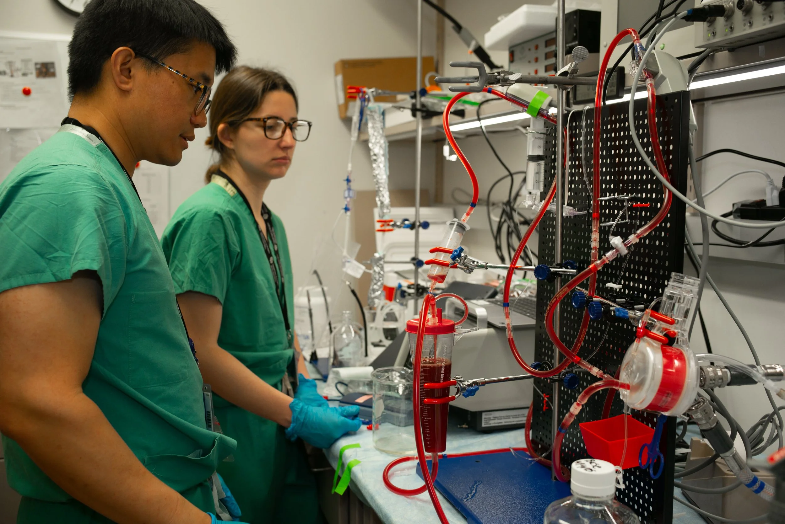 Two scientists in green scrubs working in a laboratory with various medical and scientific equipment, connected to a complex apparatus with tubes and containers.