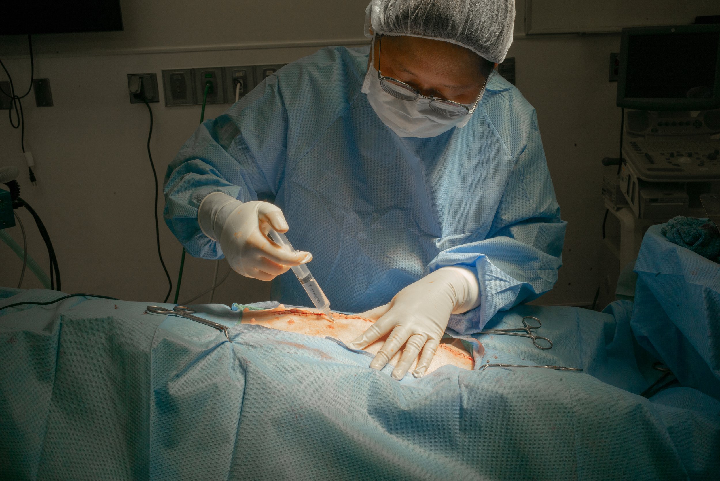 Surgeon in scrubs, mask, and goggles performing surgery on a patient, using a syringe to administer an injection or anesthesia, in an operating room.