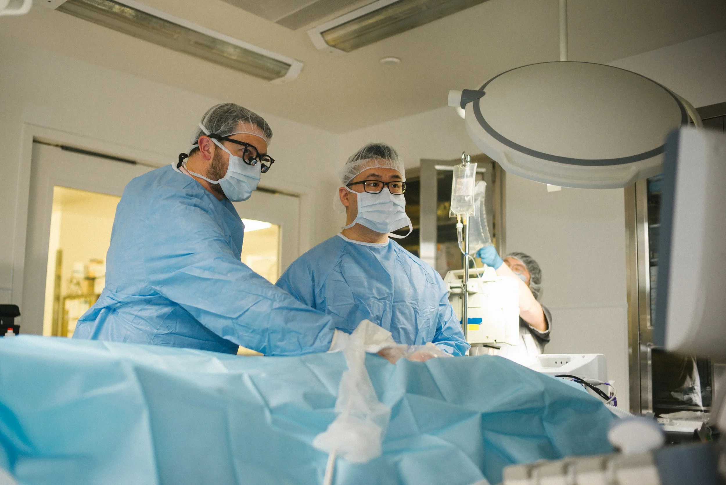 Medical professionals in surgical gowns, masks, and gloves working in an operating room with medical equipment and monitors.