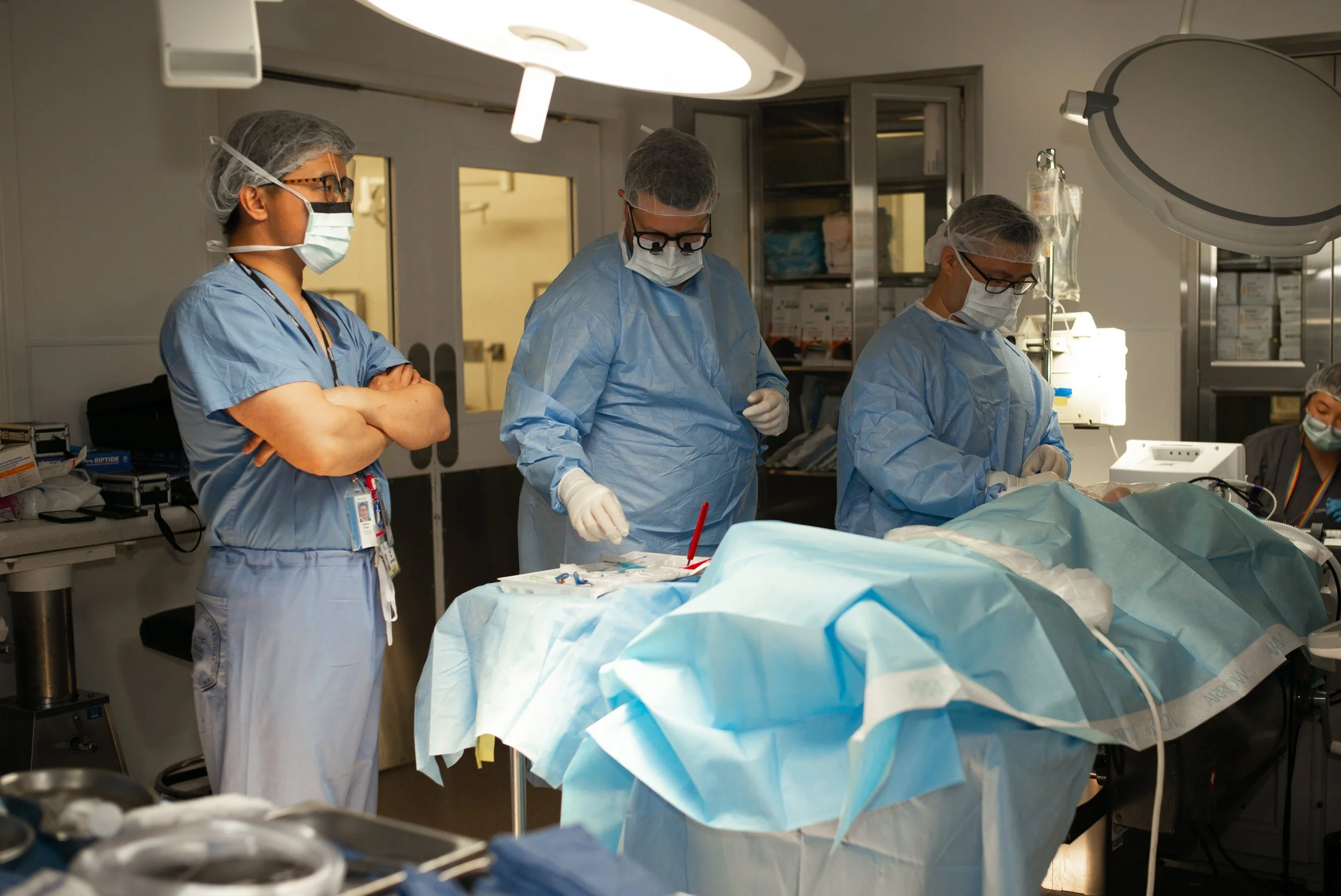 Medical team performing surgery in an operating room, wearing scrubs, masks, and gloves, with surgical instruments and equipment around a patient on the operating table.