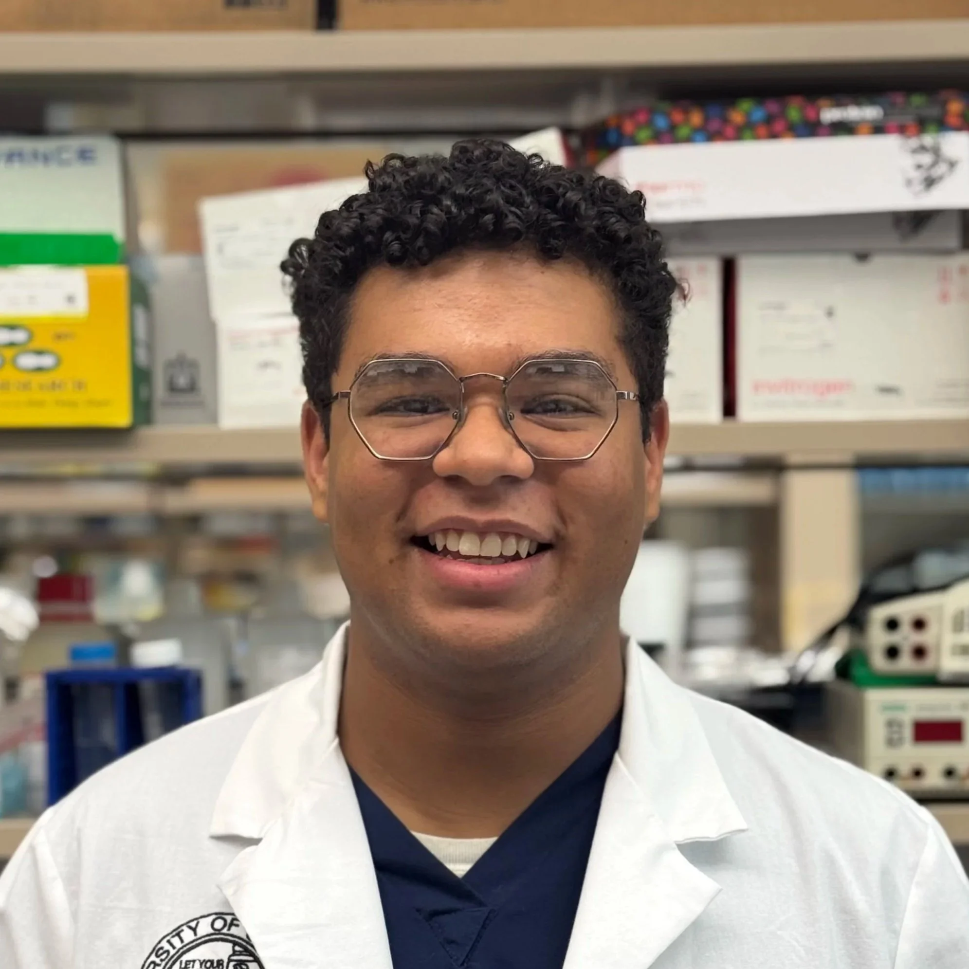 Smiling young man in a white lab coat and glasses in a pharmacy or laboratory setting.