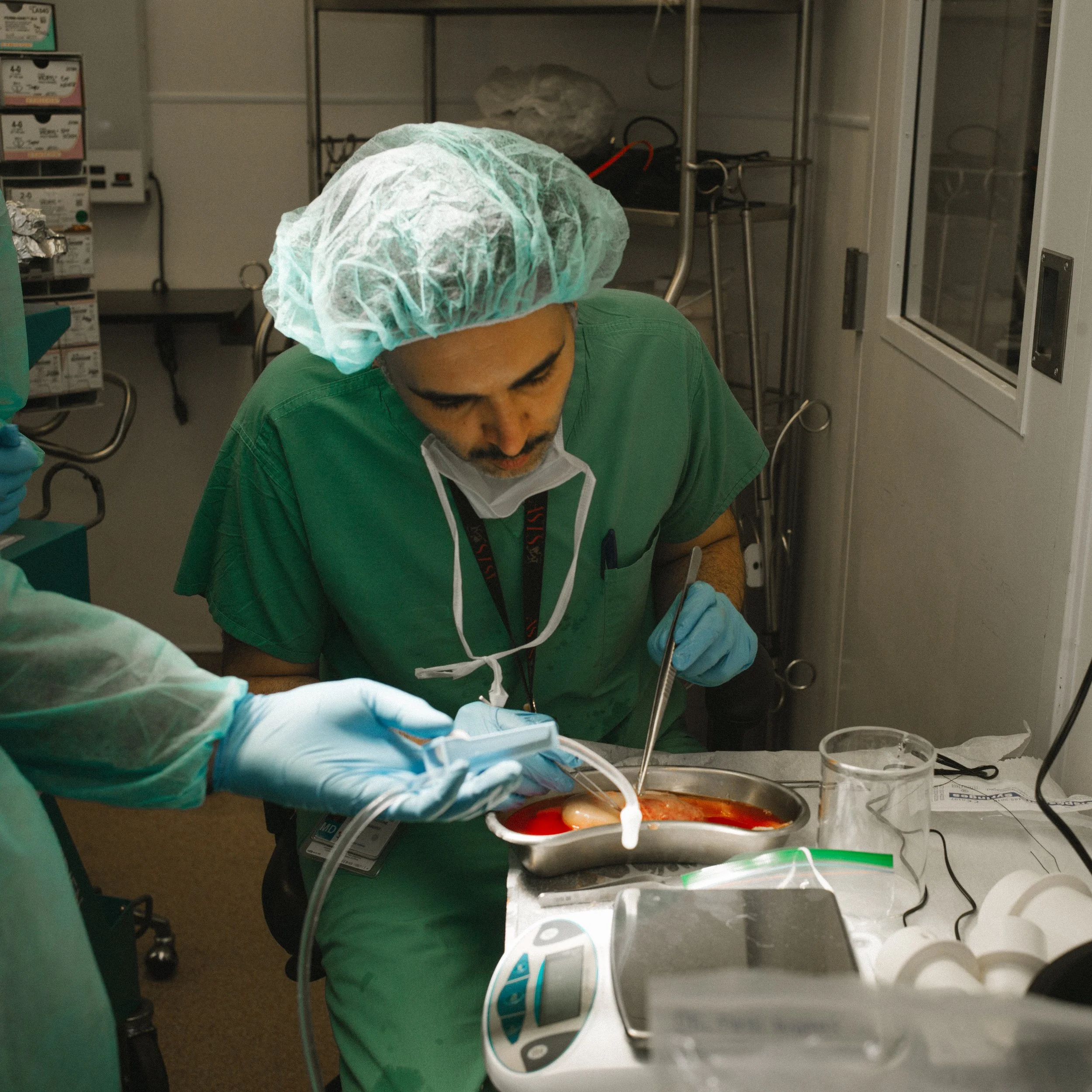 A medical professional in green scrubs, wearing a hair cover and gloves, performs a surgical procedure on a patient in an operating room.