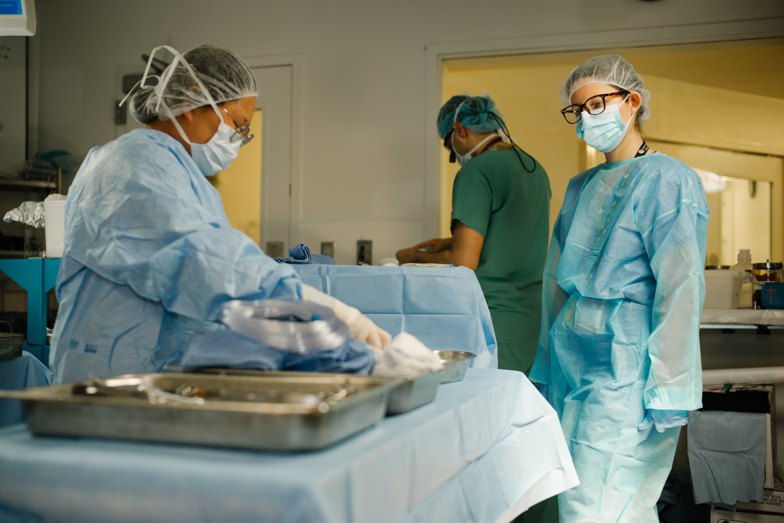 Medical professionals dressed in protective scrubs, masks, and gloves working in an operating room with surgical instruments and supplies on a table.